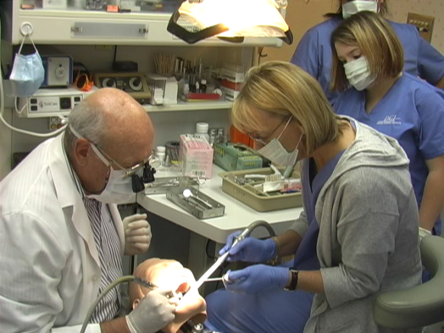 Dentist and assistant perform dental procedure with patient in dental chair, medical equipment visible in background.