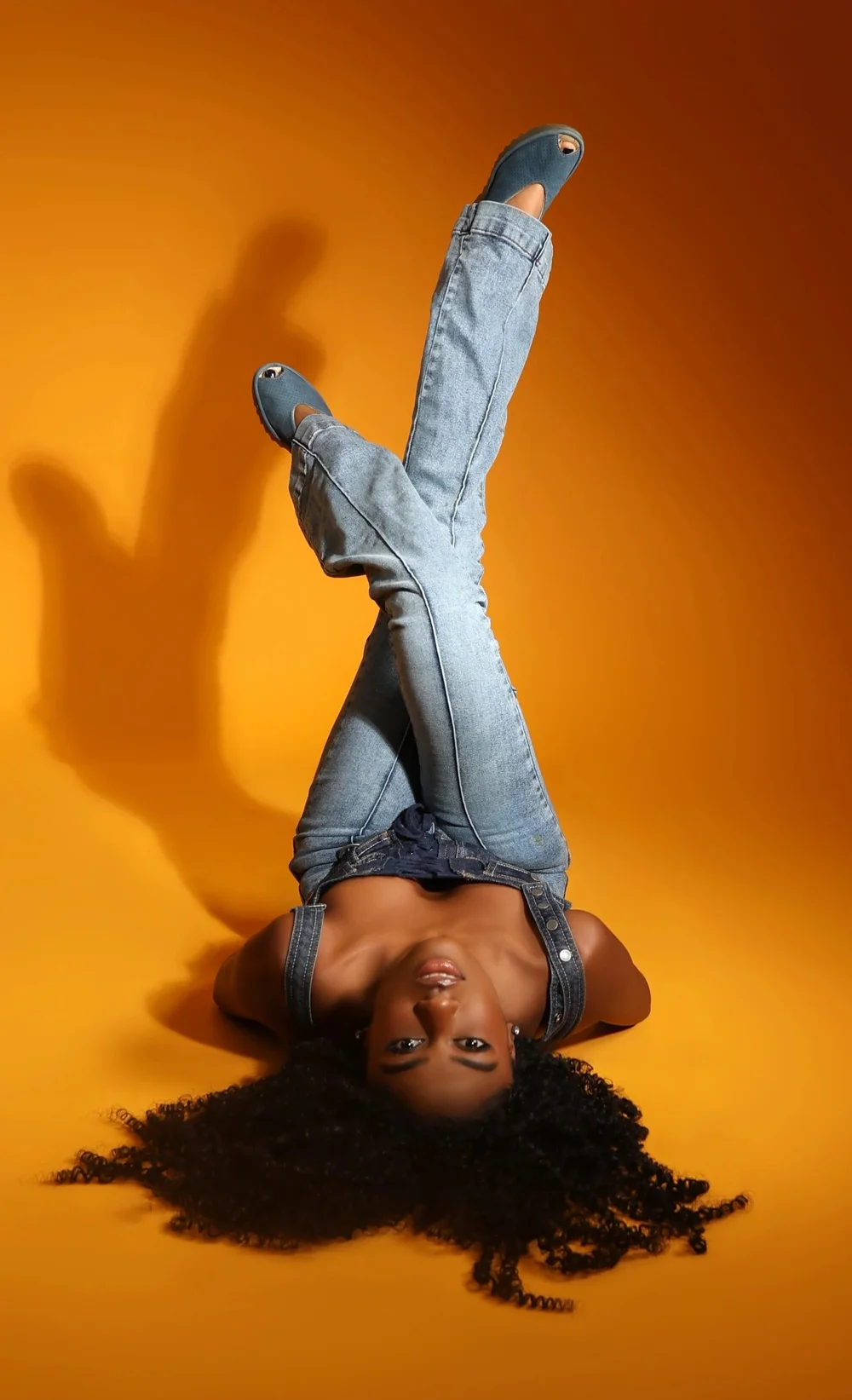 Person lying upside down with legs raised, wearing jeans and blue shoes, against an orange background.