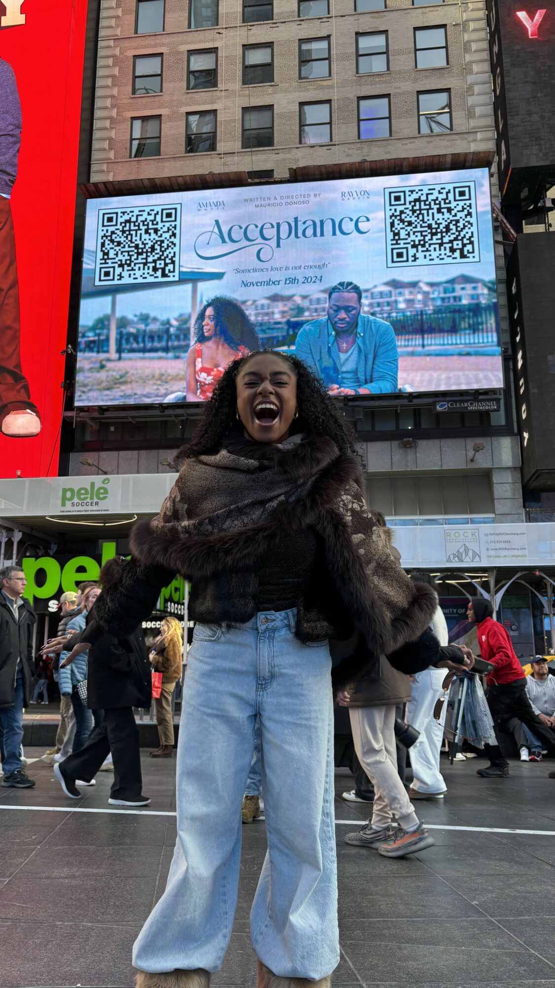 Person smiling and laughing in a busy outdoor area with a large digital billboard behind them showing two people and the word 'Acceptance'.