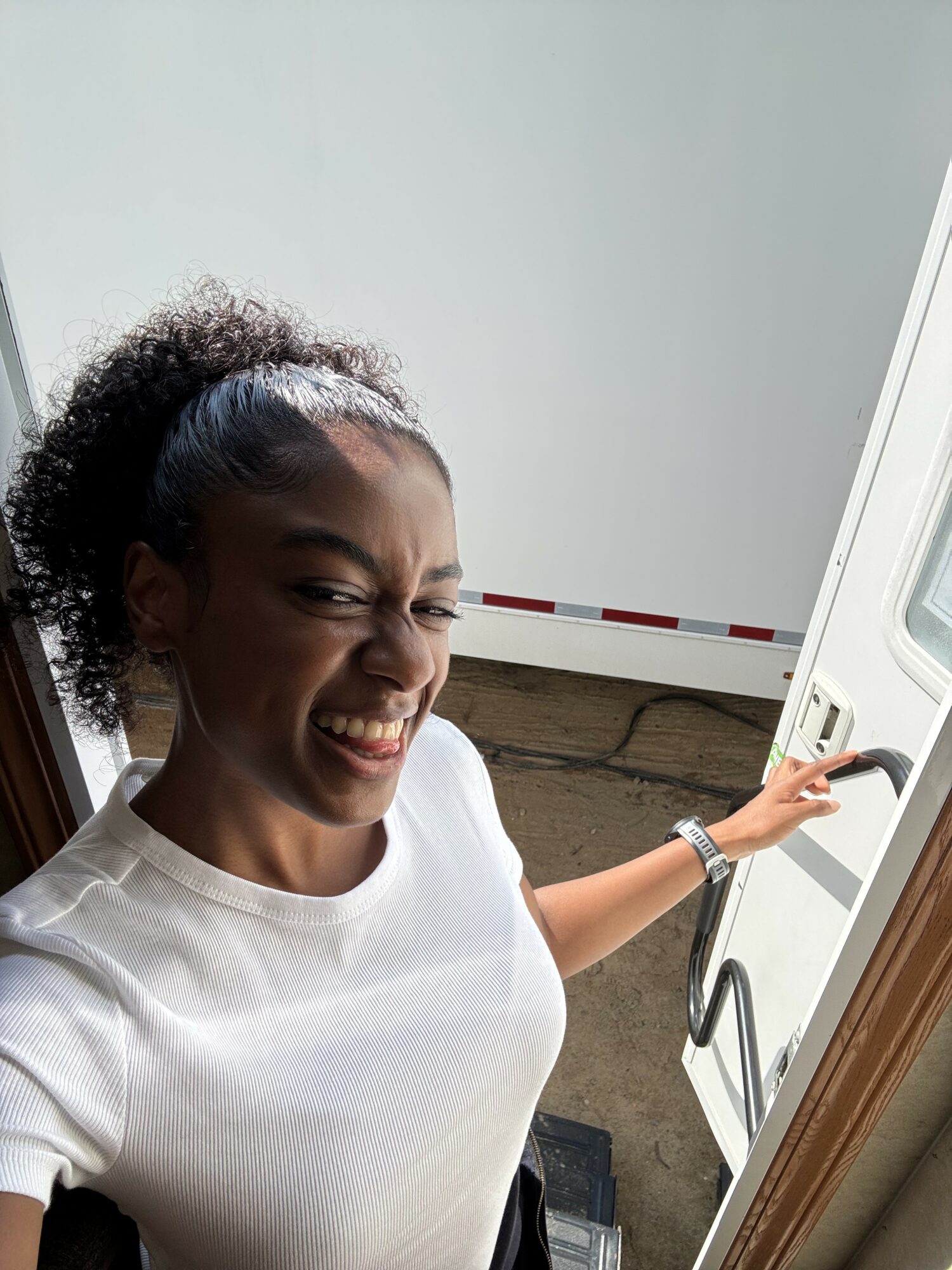Woman smiling and holding a door handle, wearing a white shirt, with a neutral background.