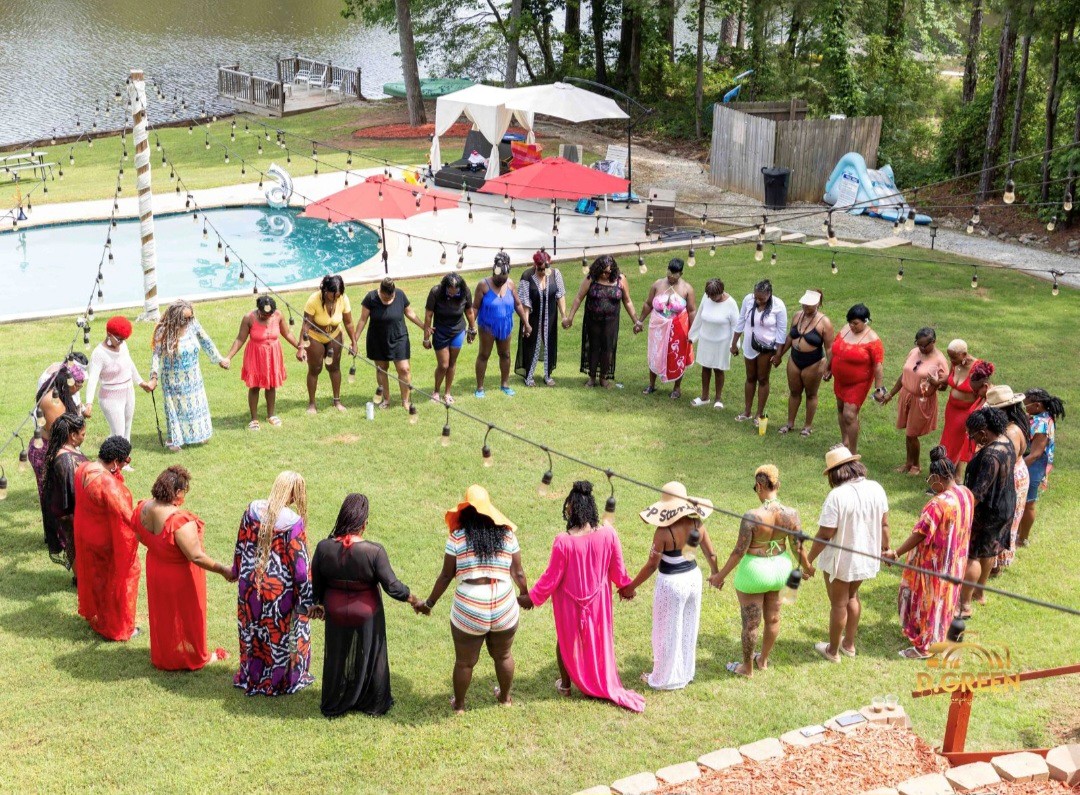 Group of people holding hands in a circle on a grassy area outdoors, with pool and trees in background.