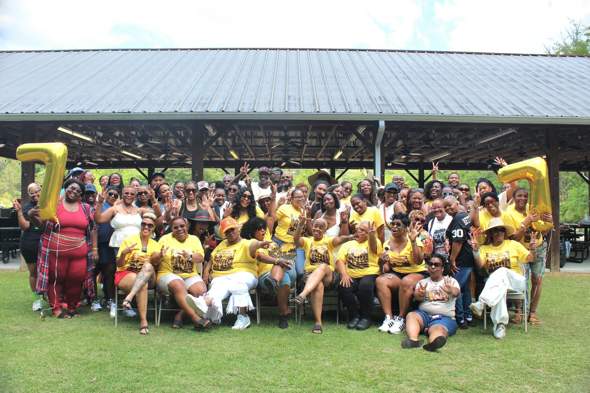 Group of people gathered outdoors under a pavilion, some wearing yellow shirts, on grass with trees in background.