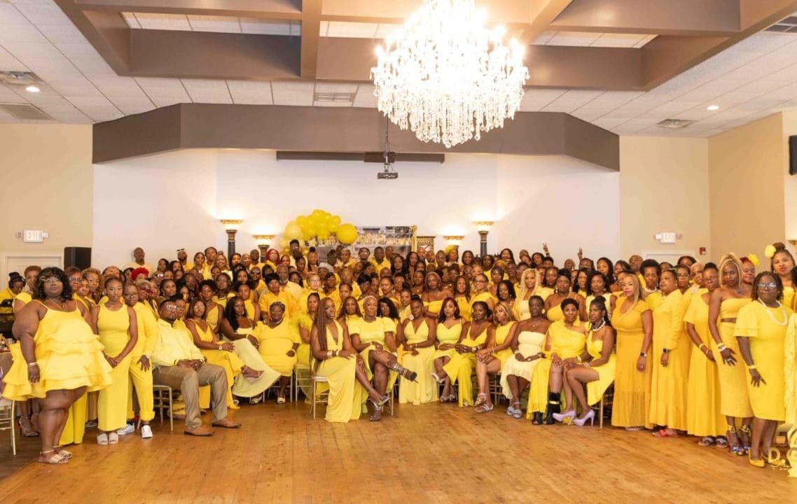 Large group of people in yellow clothing gathered in a banquet hall with a chandelier and floral decorations.