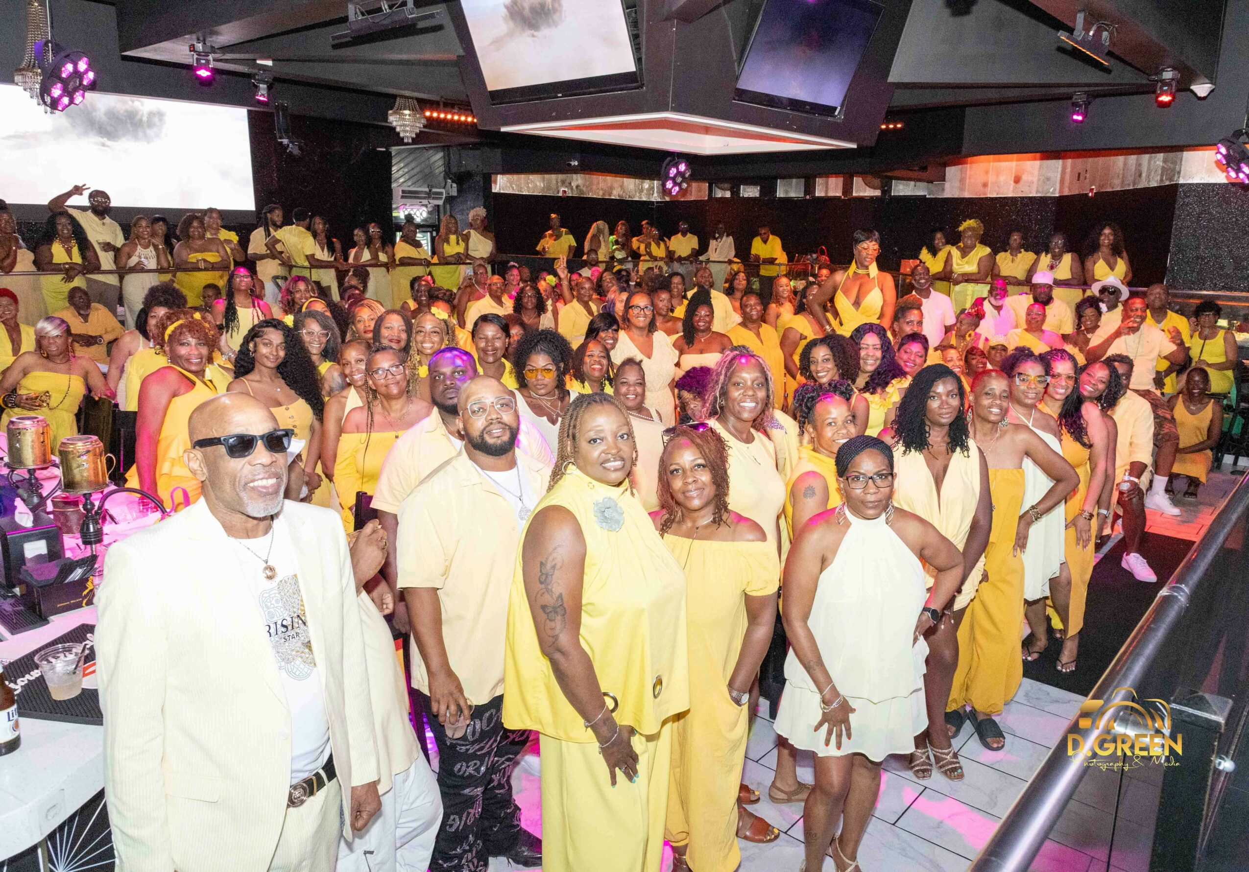 Group of people in yellow and white outfits gathered in a decorated indoor event space with tables and ceiling screens.