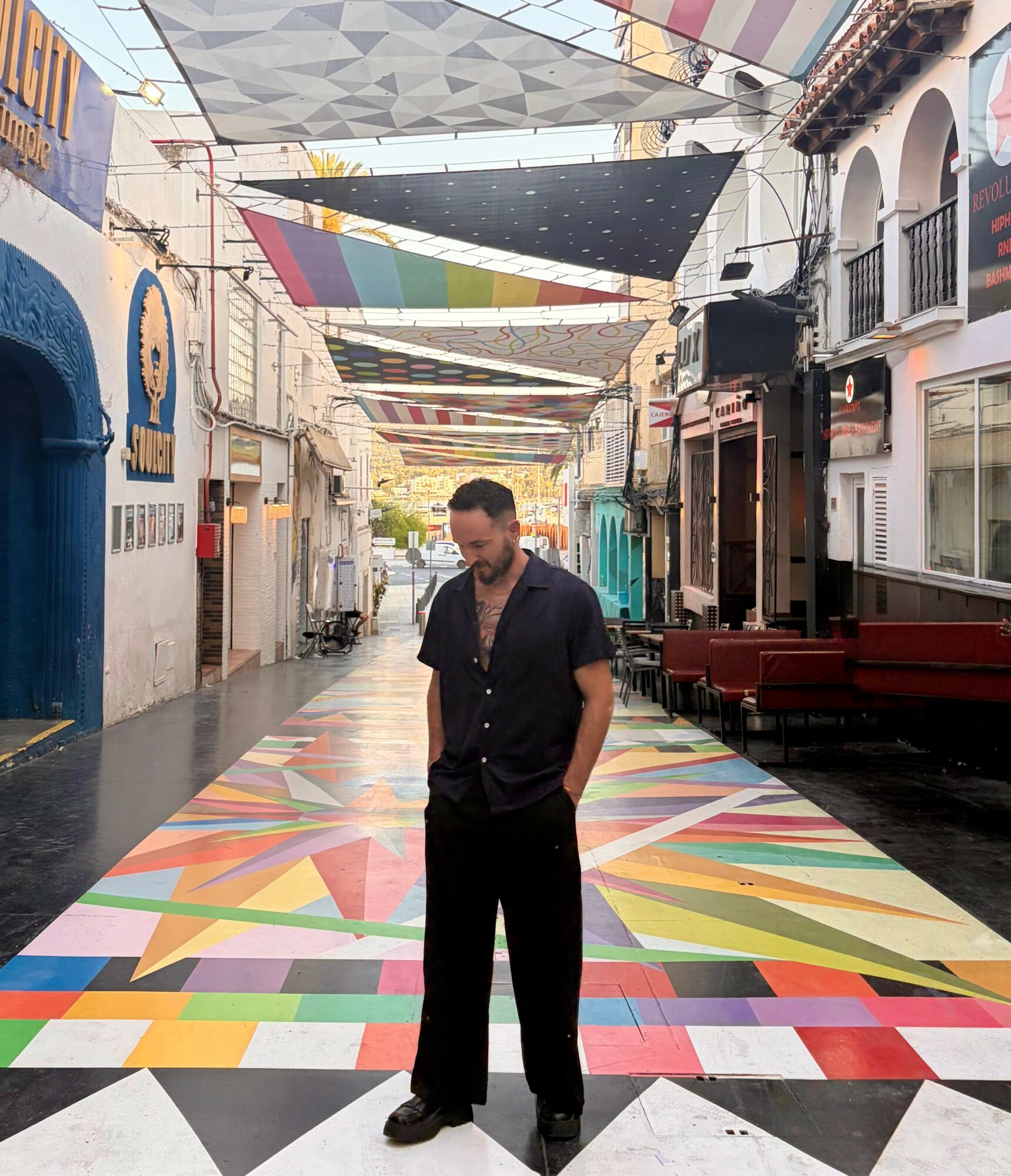 Man standing on colorful patterned street with buildings and decorative overhead structures, looking down with hands in pockets.