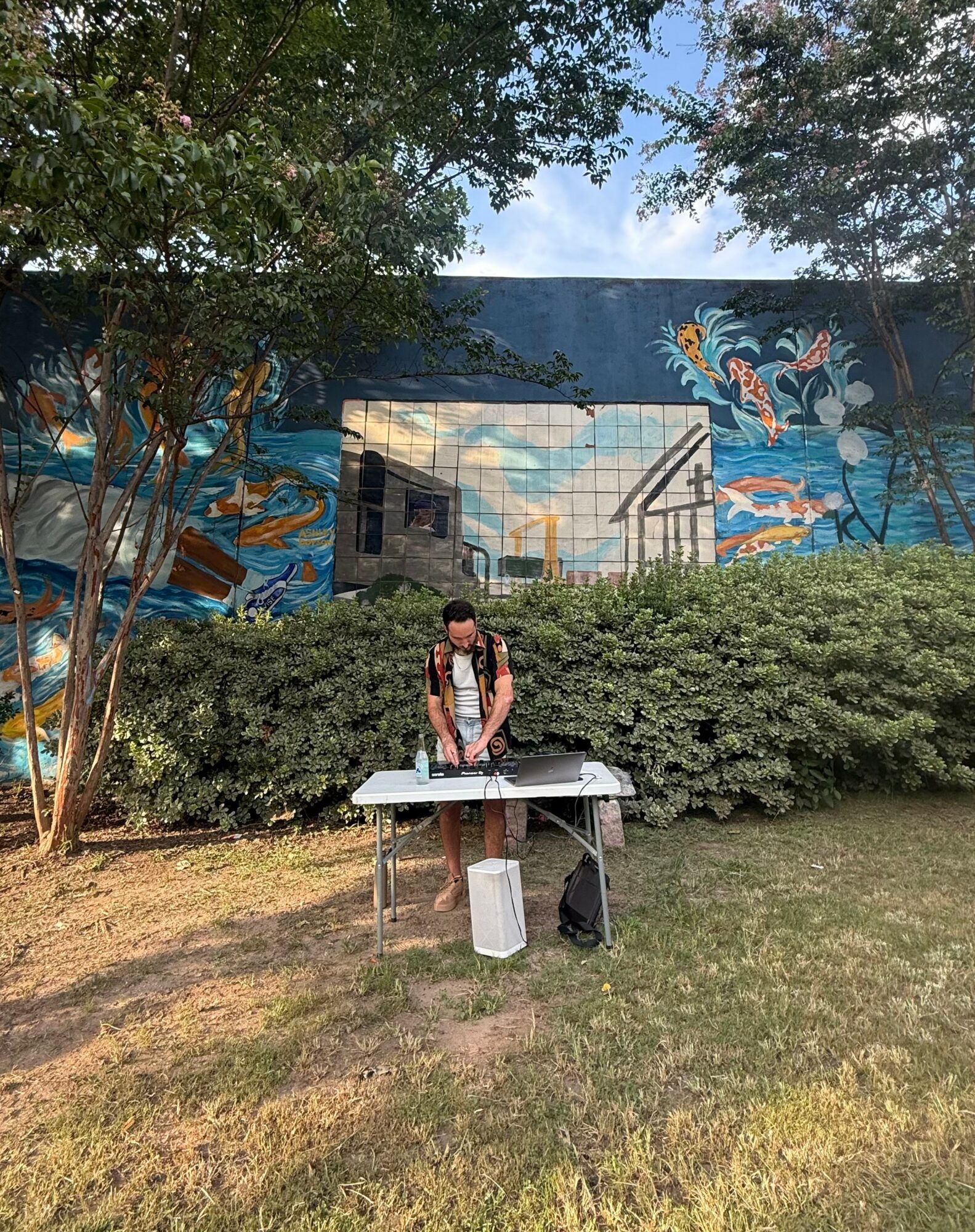 Person standing at a table outdoors with a mural and trees in the background.