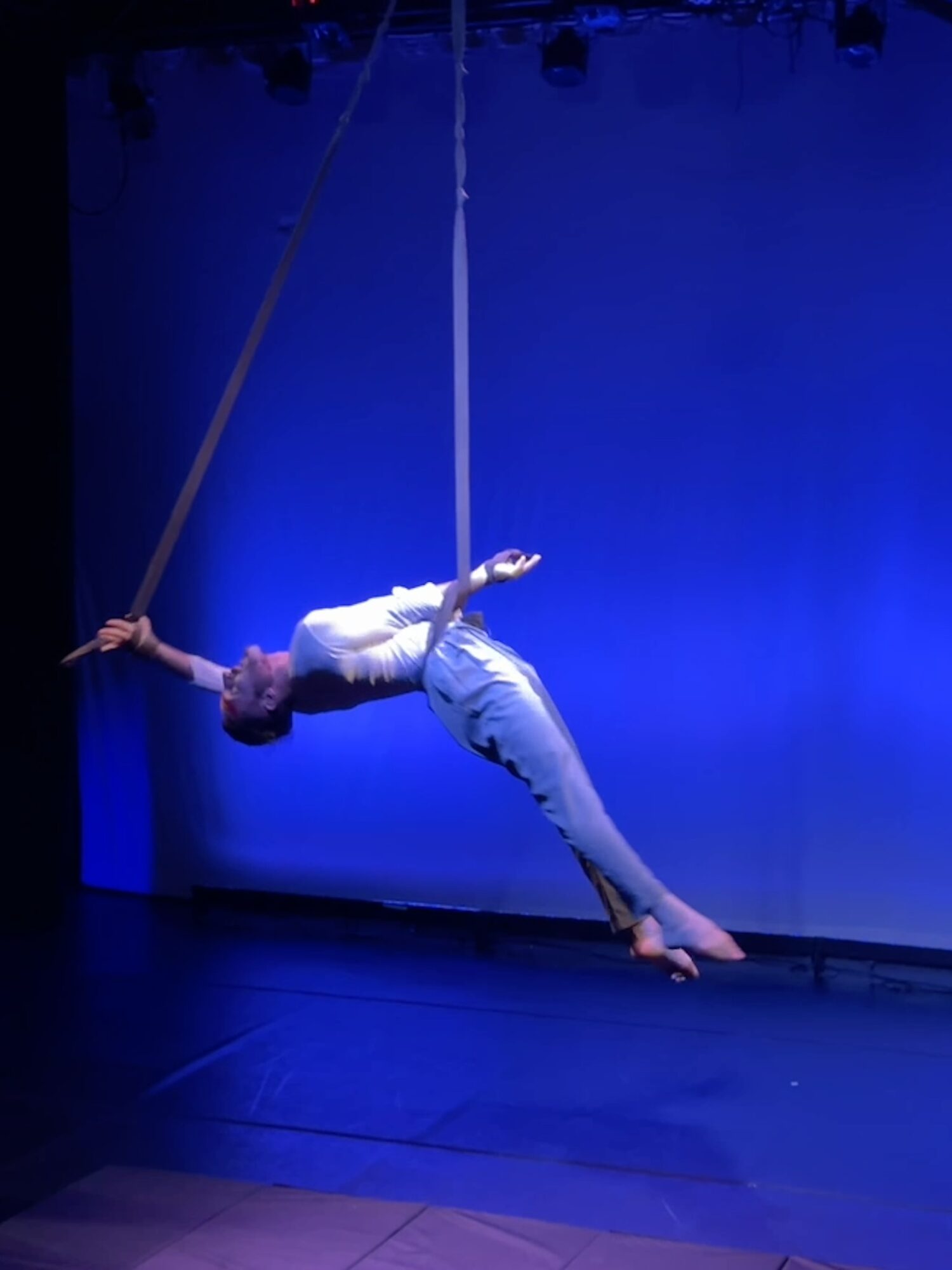 Performer hanging horizontally on aerial silk against blue background, with arms extended and legs bent, in a dance or acrobatic pose.