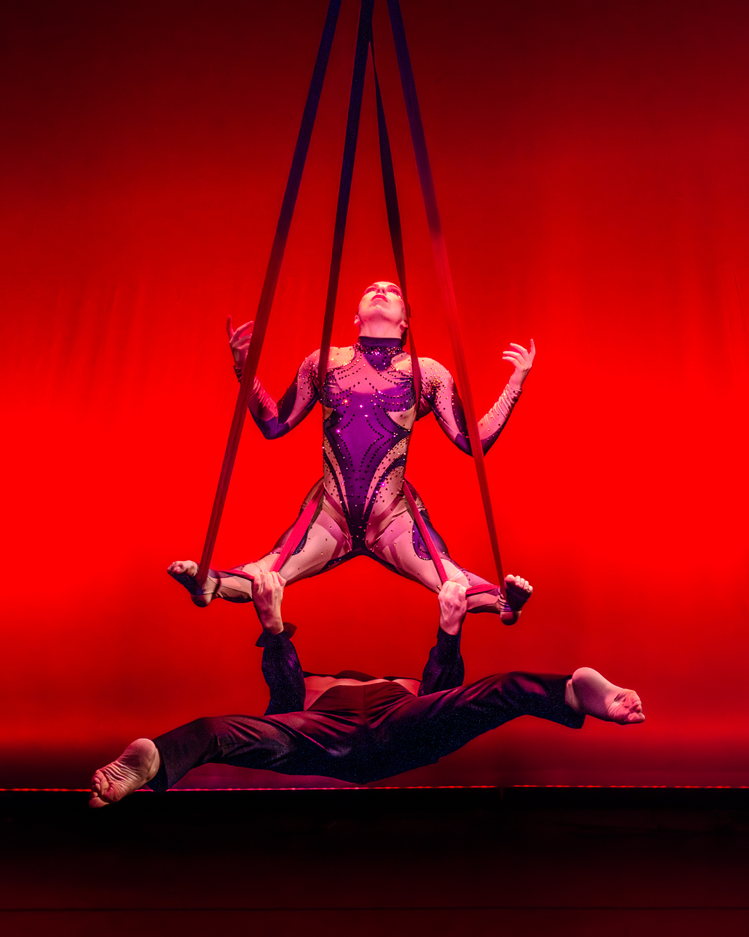 Aerial performer in purple costume on red background, supported by two people lying on stage, with ropes overhead.
