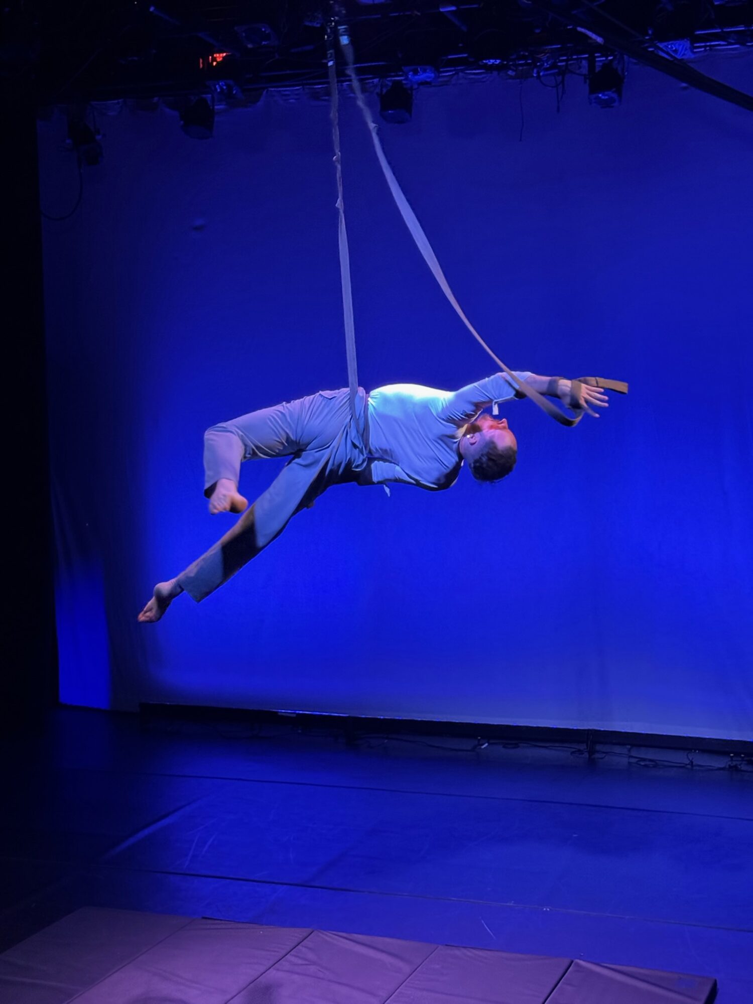 A person performs aerial silk acrobatics on stage with a blue background, hanging horizontally with one arm extended.