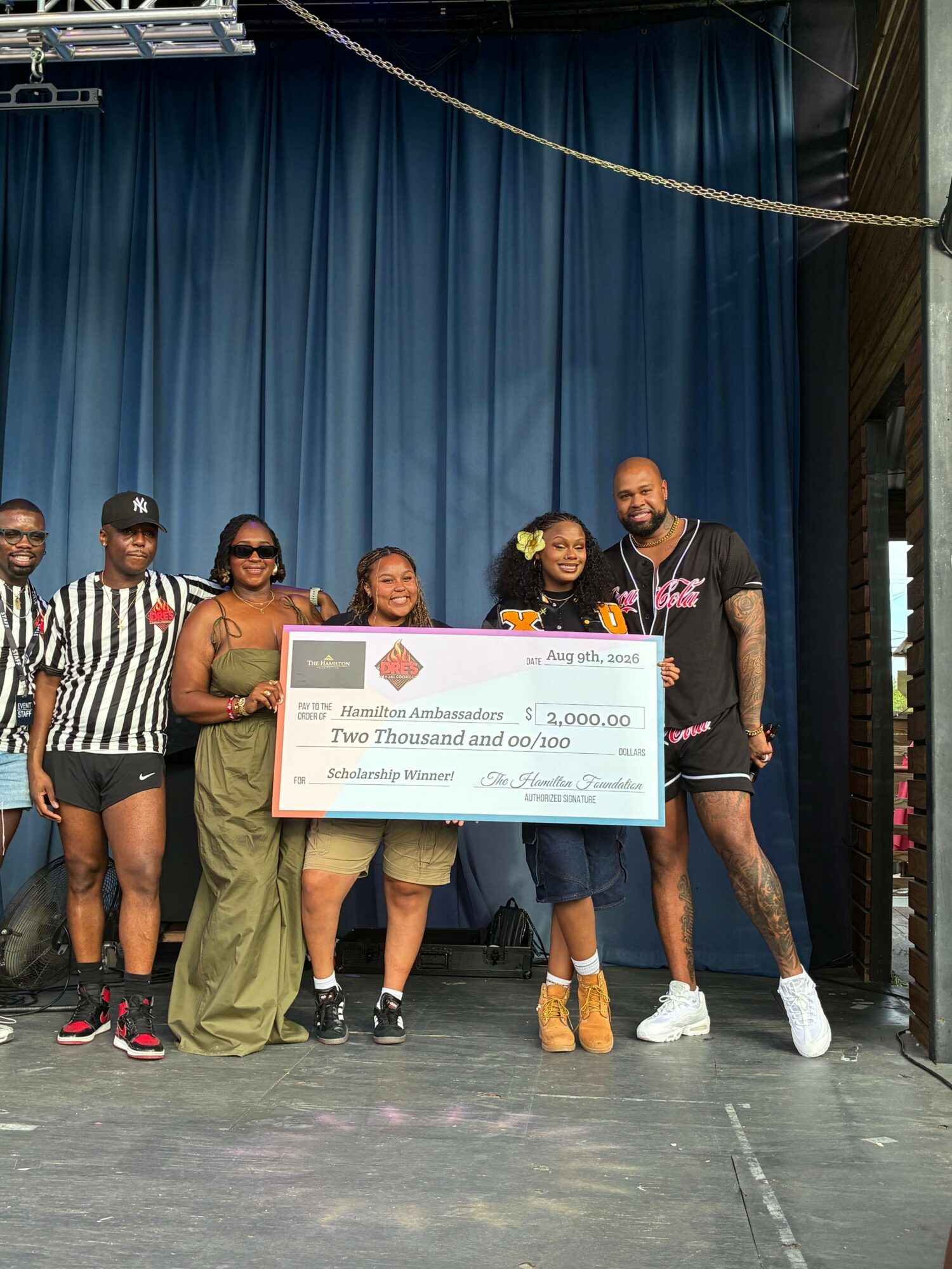 Four people on stage holding a large check, with a blue curtain background and a person in a striped shirt.