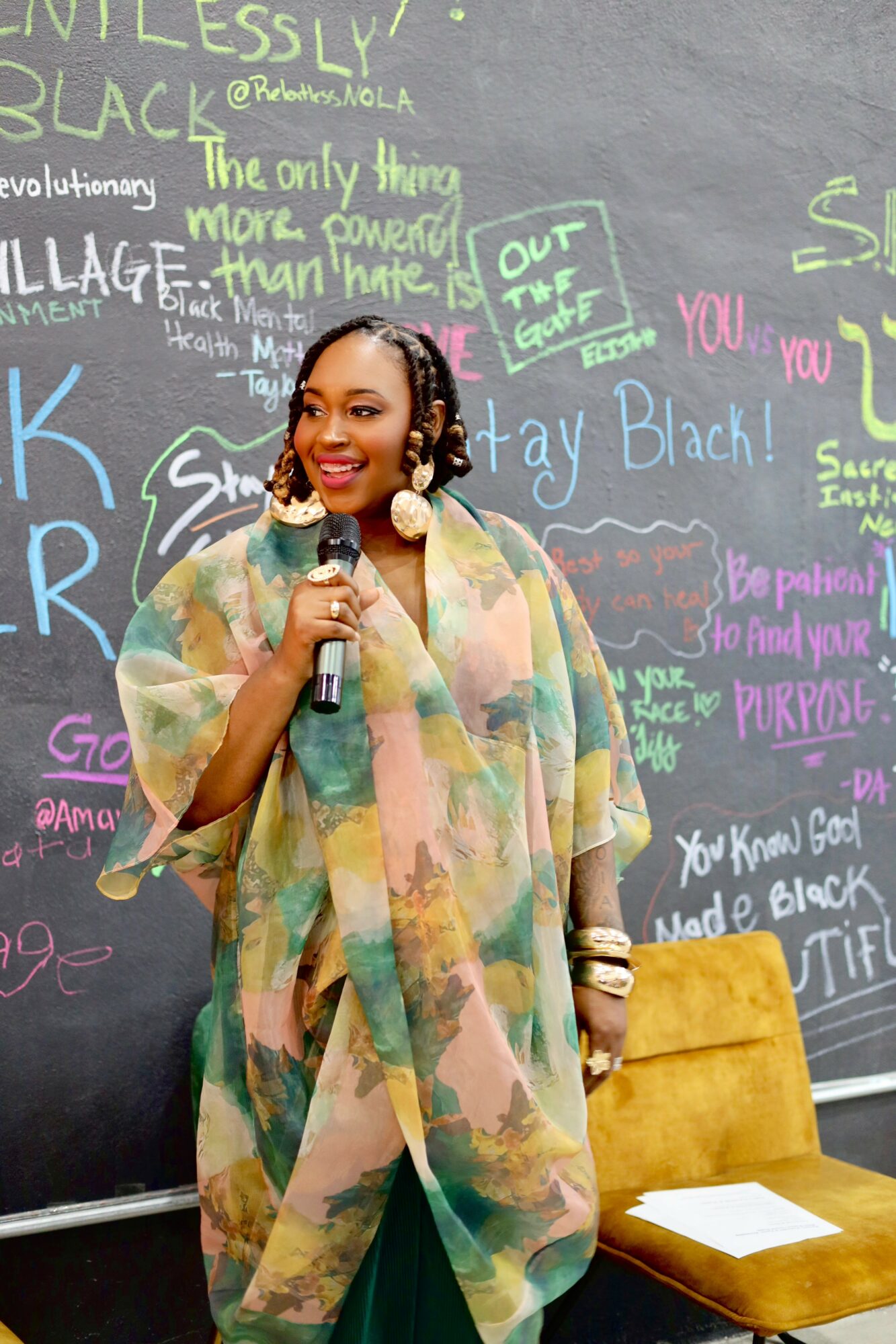 Woman with short hair and earrings holding a microphone, standing in front of a colorful chalkboard with messages and drawings.