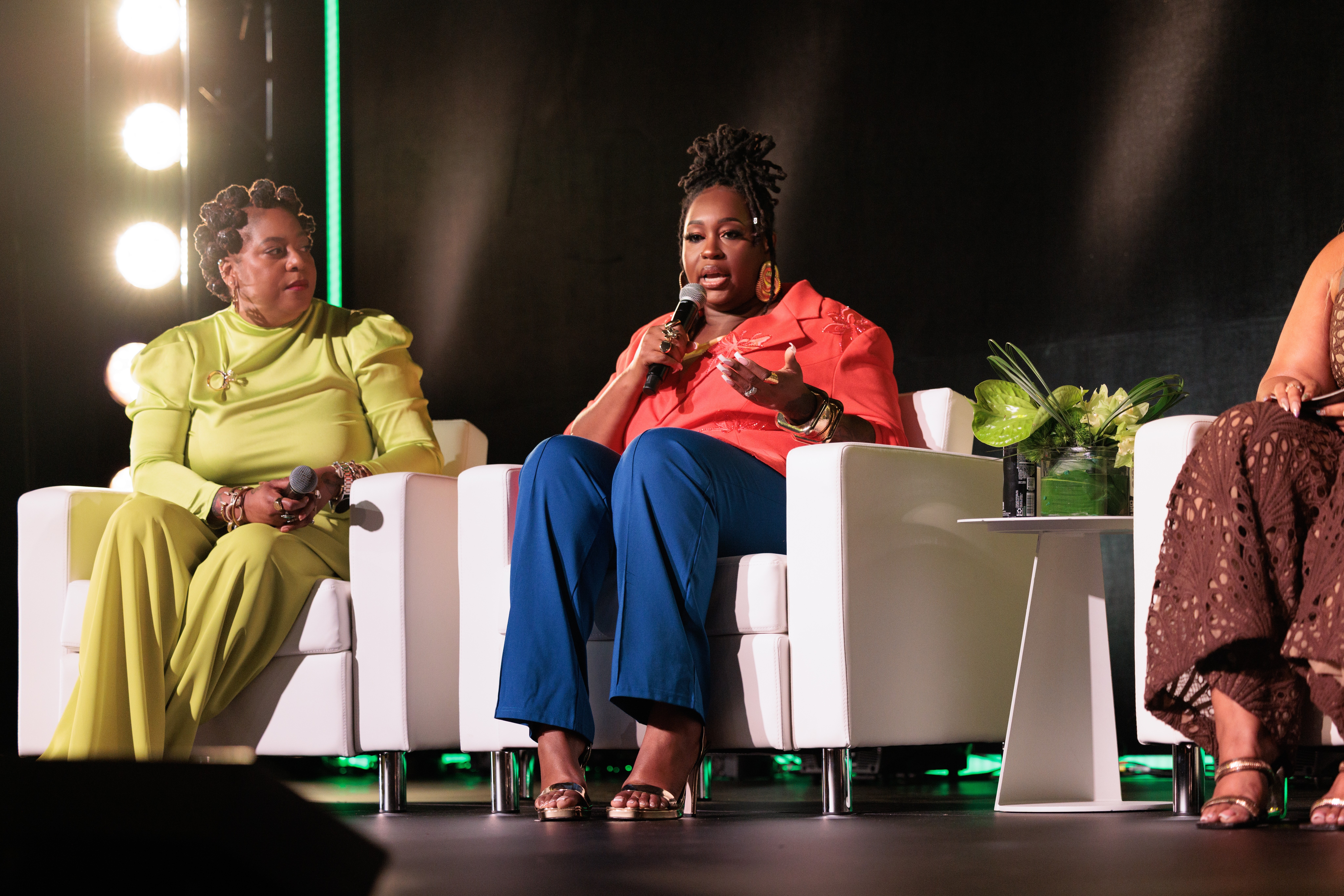 Three women seated on stage, one speaking into a microphone, with plants and a black background.