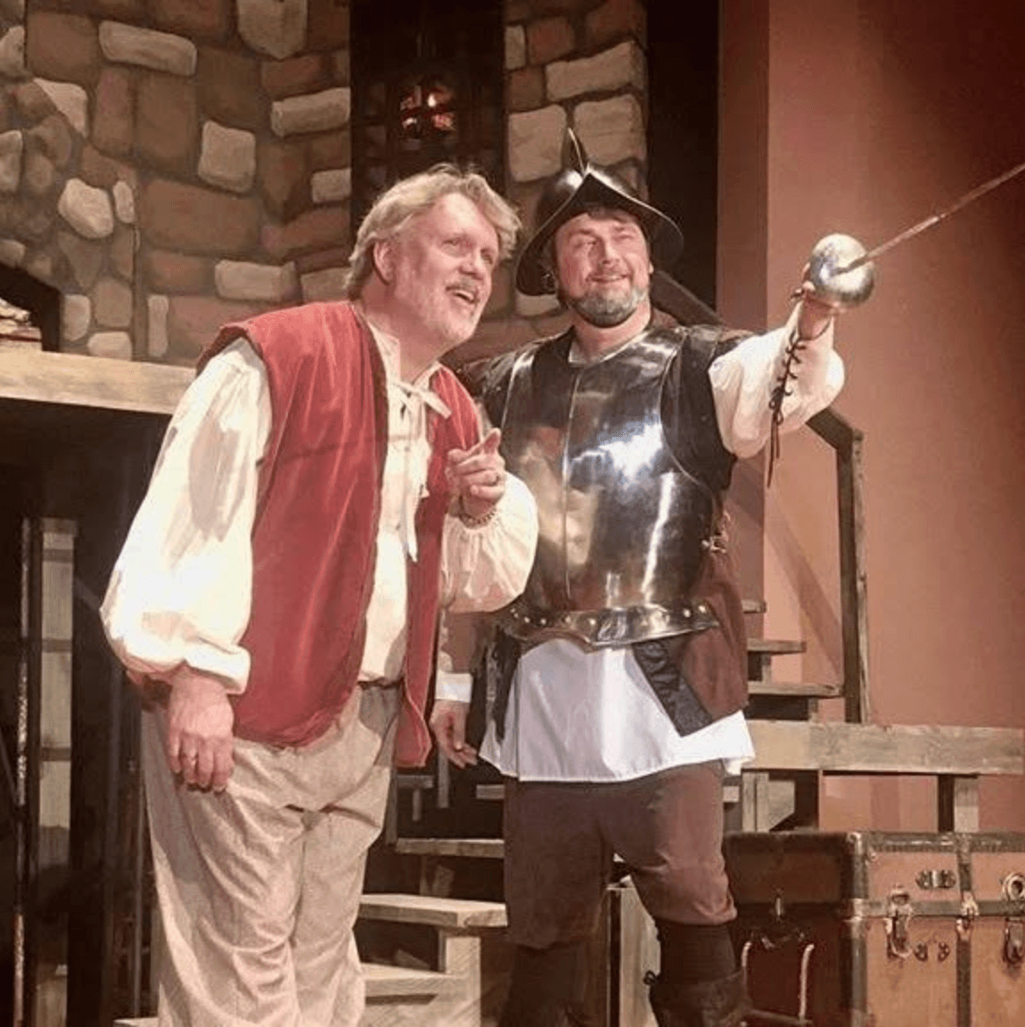 Two men in costume, one smiling and pointing, the other dressed as a pirate, standing indoors with a stone wall background.