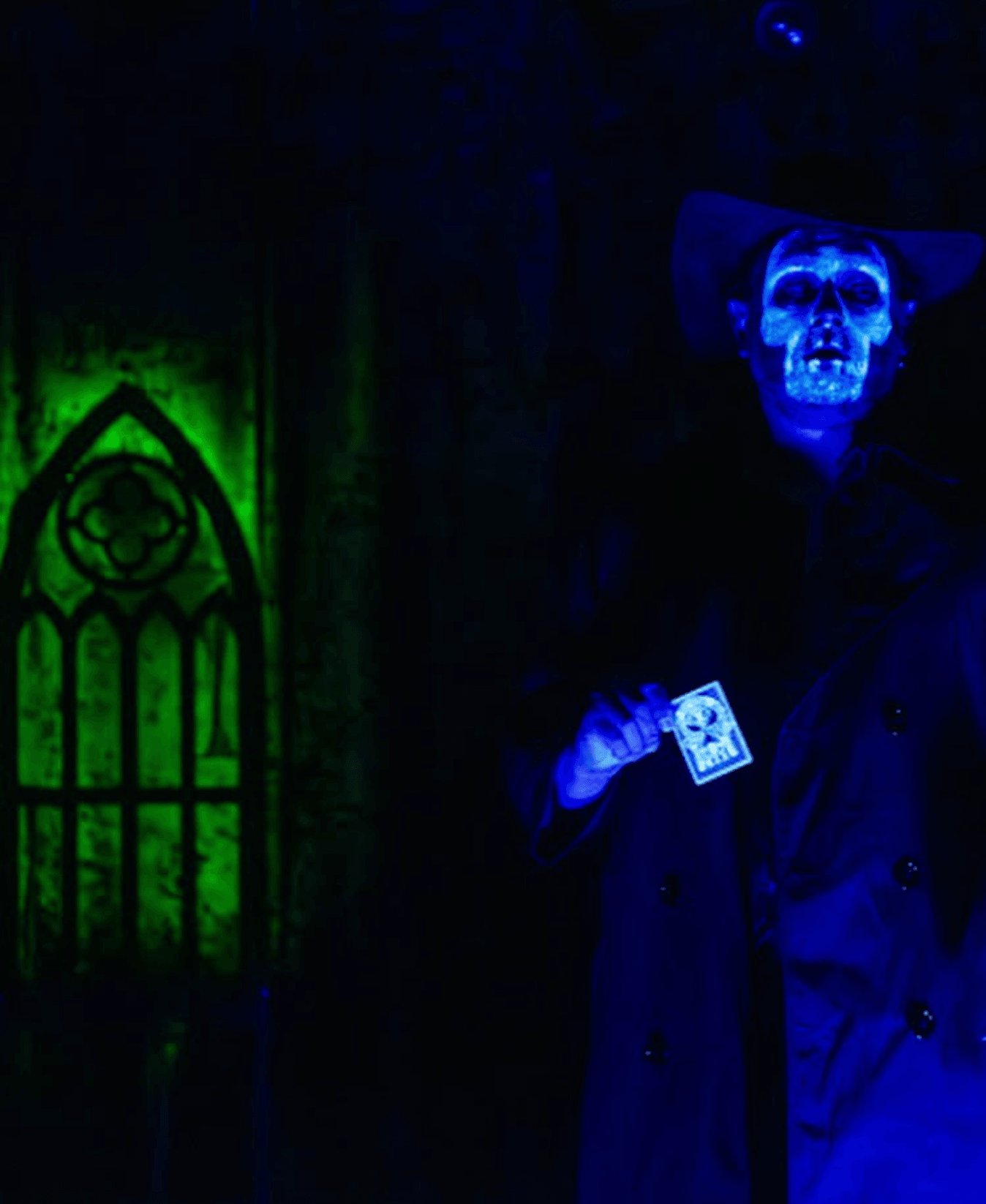 Person wearing a glowing blue skull mask holding a card, standing in front of a green-lit gothic window, dark background.
