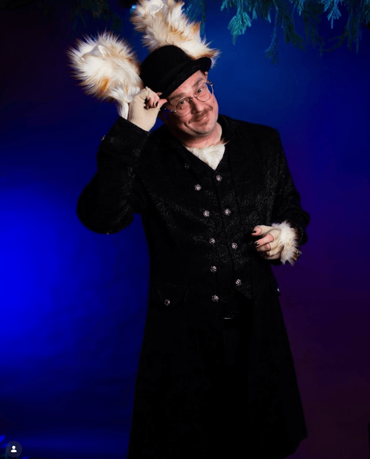 Person wearing a black coat and hat, holding furry ears, smiling, against a blue background with green foliage.