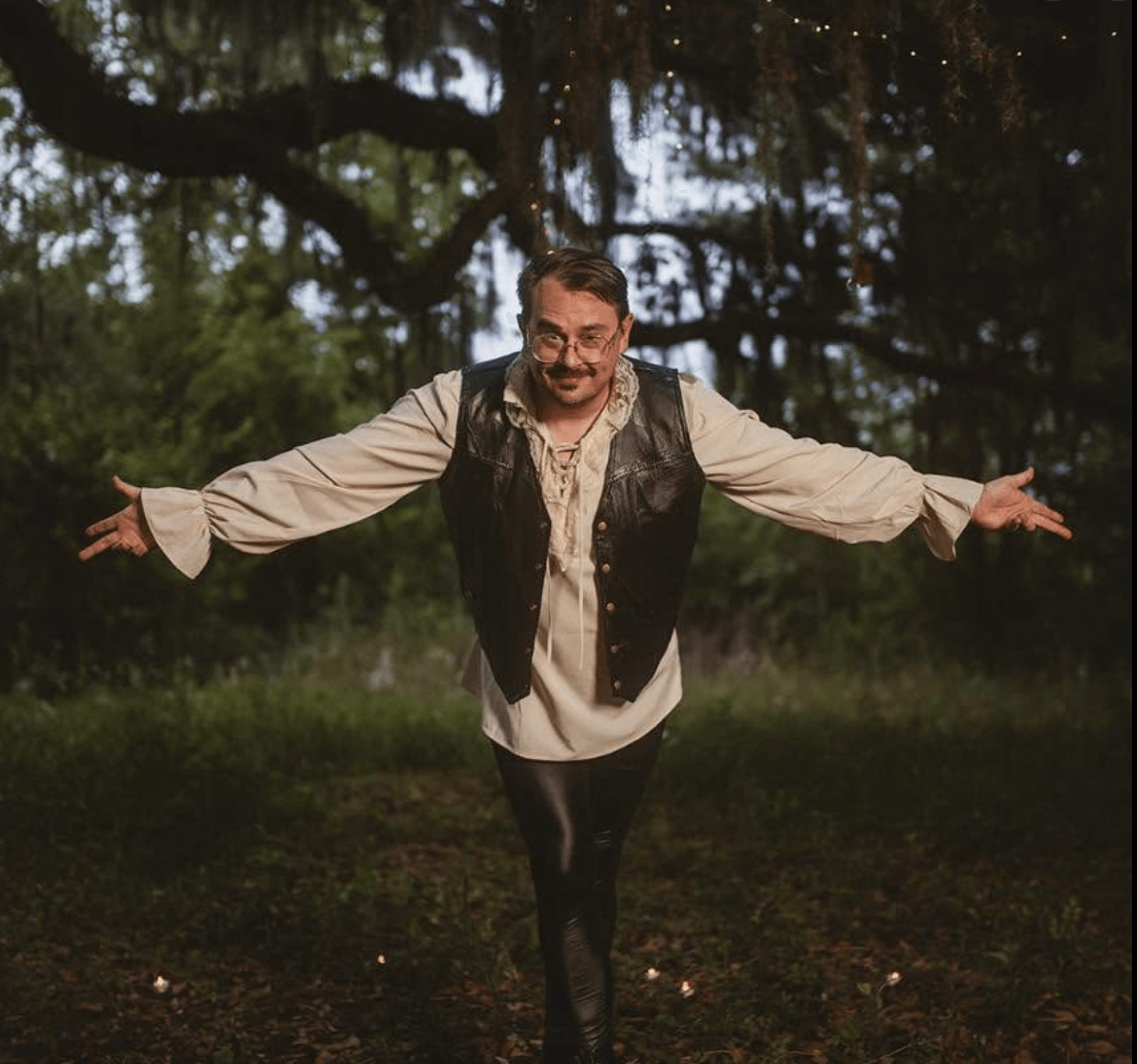 Person standing outdoors with arms outstretched, smiling, in a wooded area with hanging moss, during daytime.