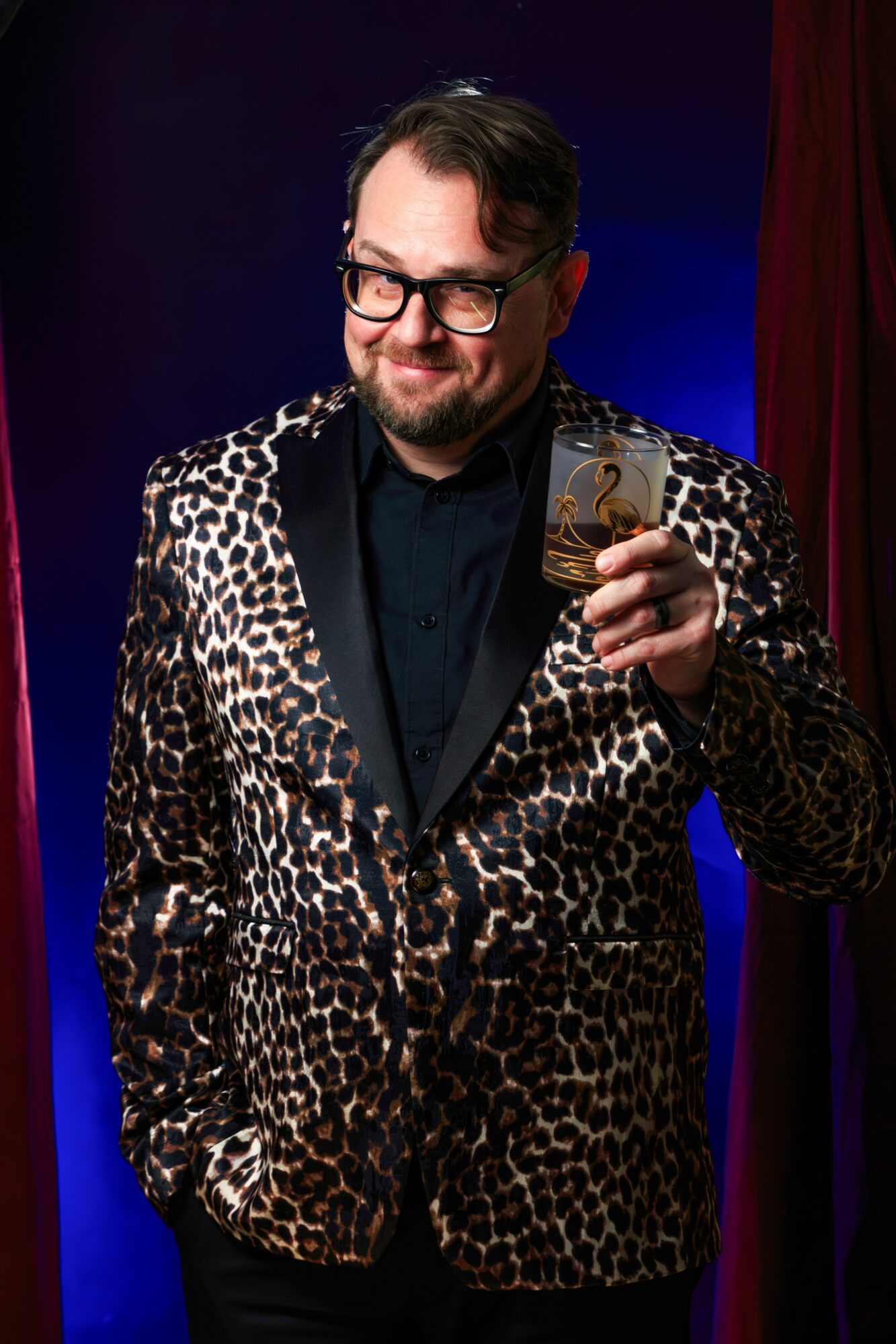 Man with glasses and a beard in a leopard print blazer holding a card, smiling, against a dark background.