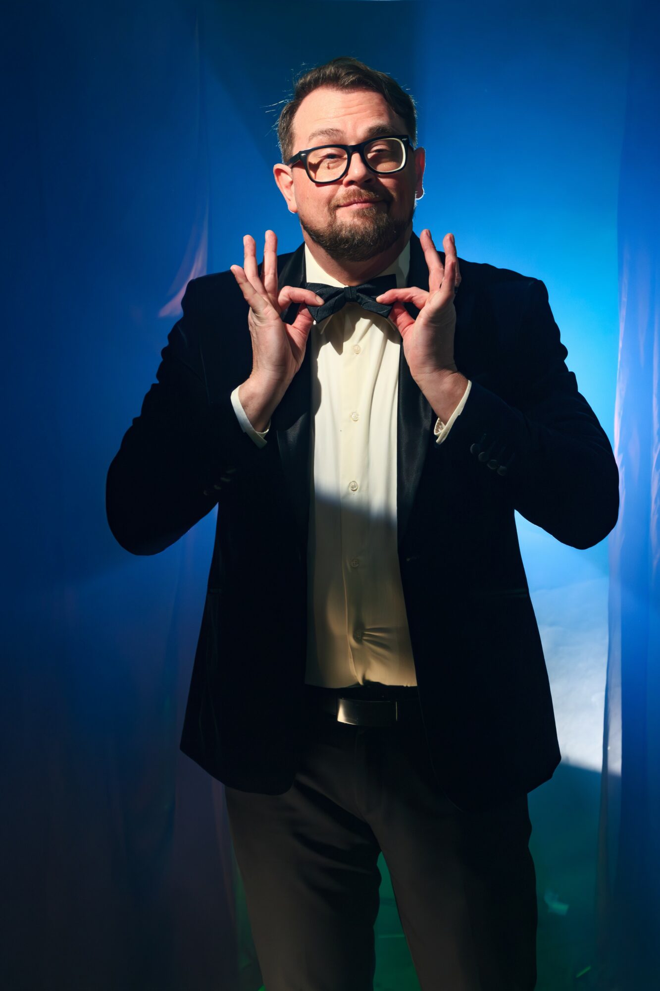Man with glasses and beard in tuxedo making a gesture with both hands, standing against a blue background.
