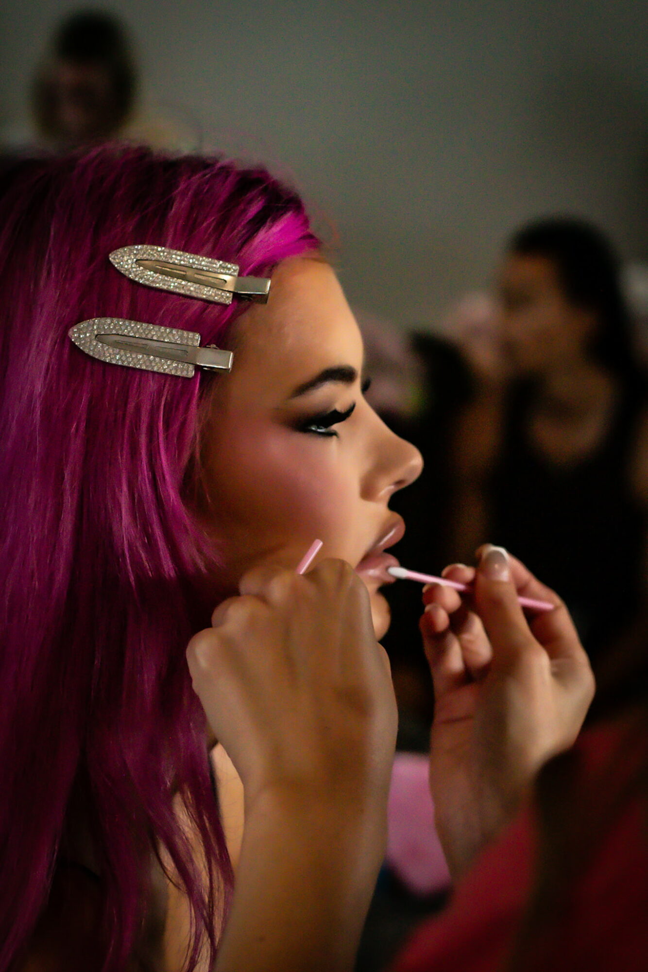 Woman with pink hair getting makeup applied, with blurred people in background.