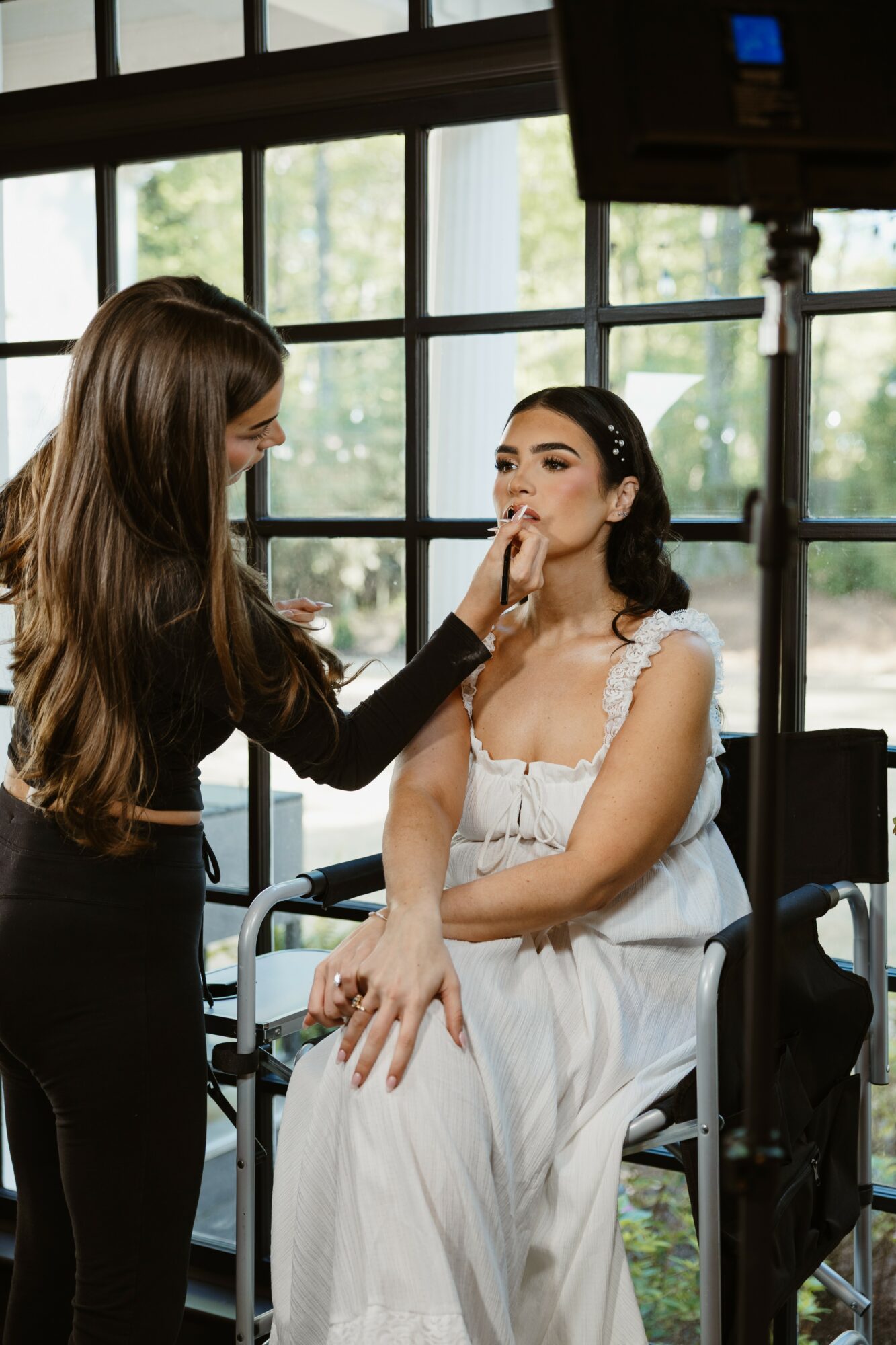 Makeup artist applies makeup to a woman sitting on a chair in front of large windows, natural light coming in.