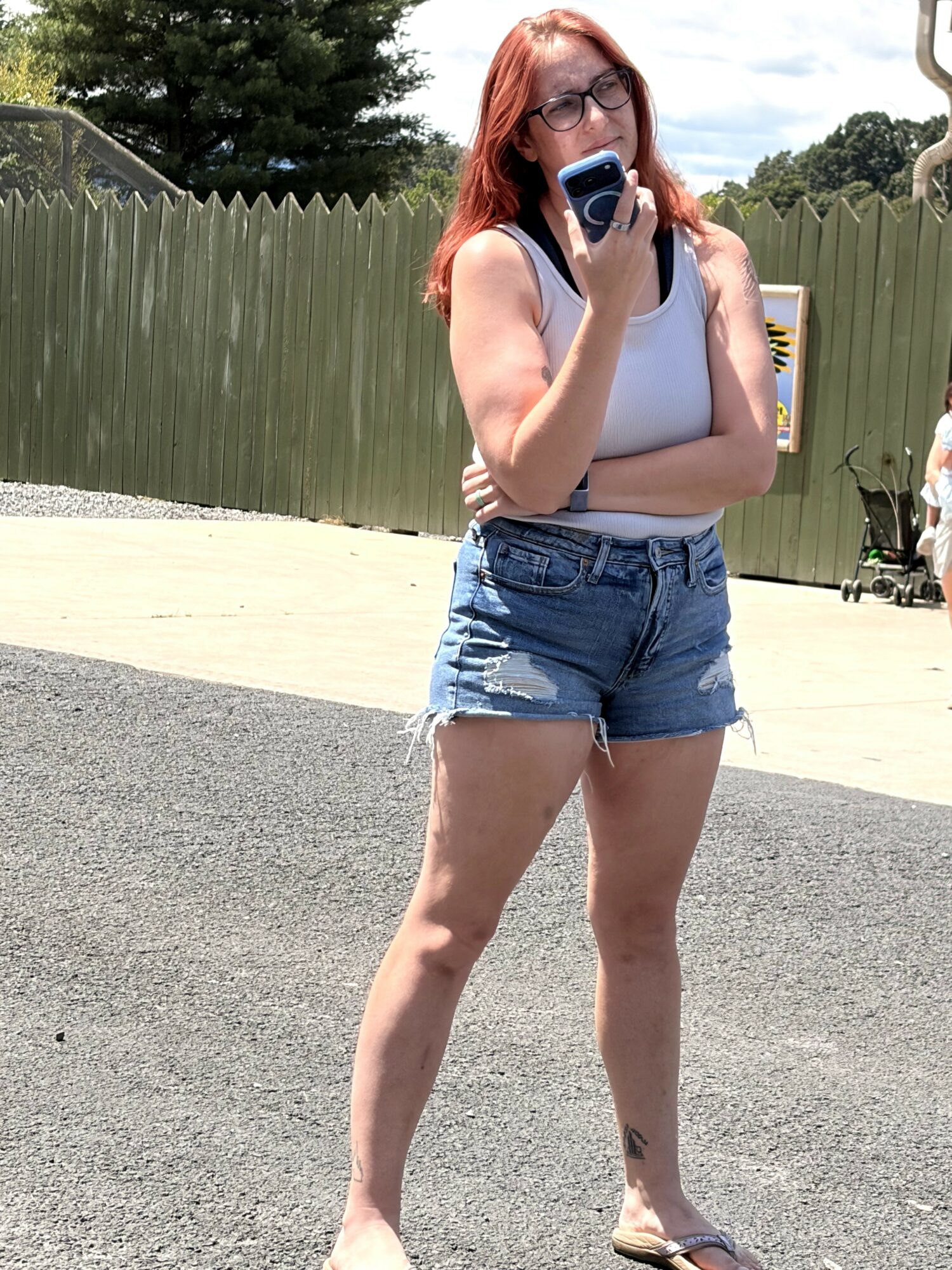 Woman with red hair and glasses stands outdoors, holding a phone to her mouth, wearing a white tank top and denim shorts.
