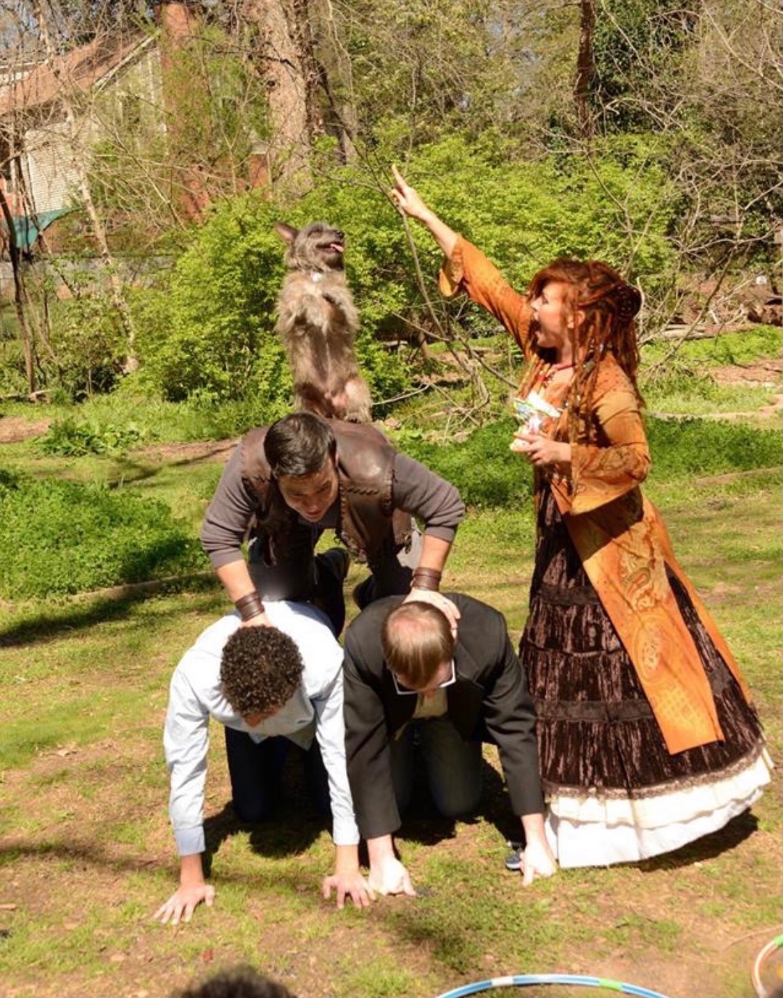 Four people in a grassy yard with trees, two kneeling and two standing, with a dog in the background. One person is raising an arm.