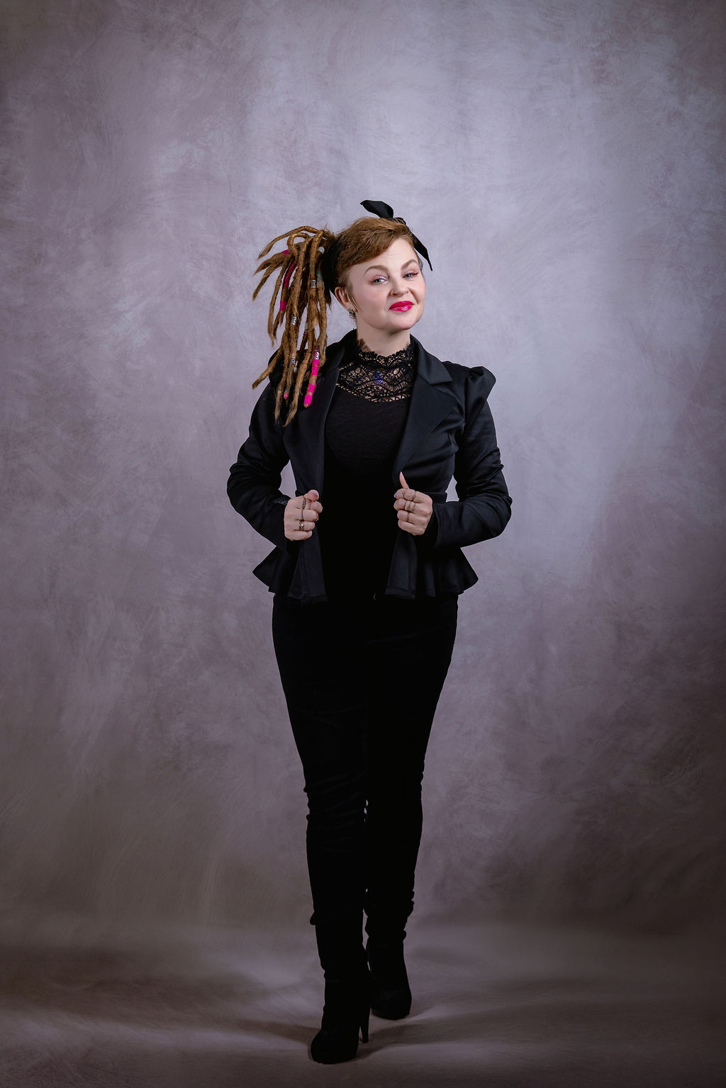 Woman with dreadlocks wearing a black jacket and pants, standing against a gray background.
