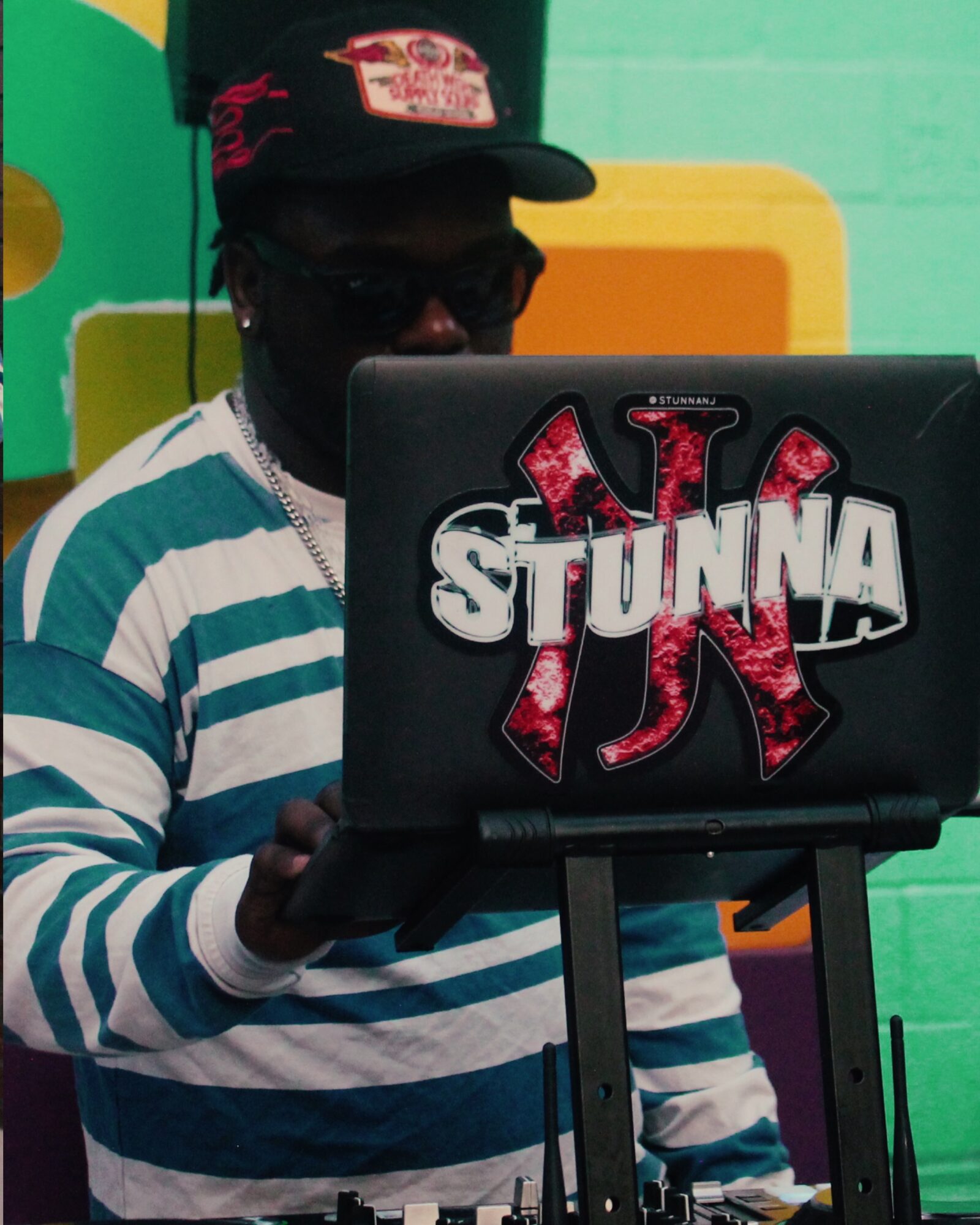 Person wearing a striped shirt, cap, and sunglasses using a laptop with a sticker that says 'STUNNA'.