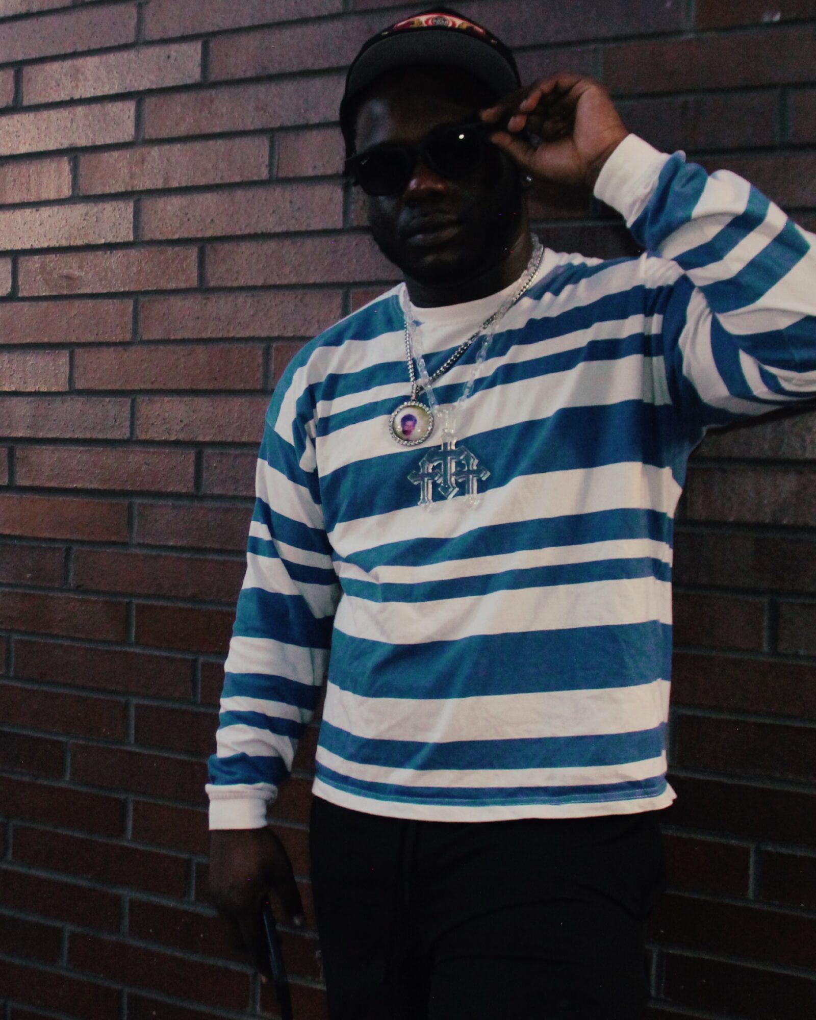 Person wearing sunglasses and a striped blue and white shirt, standing against a brick wall, adjusting their hat.