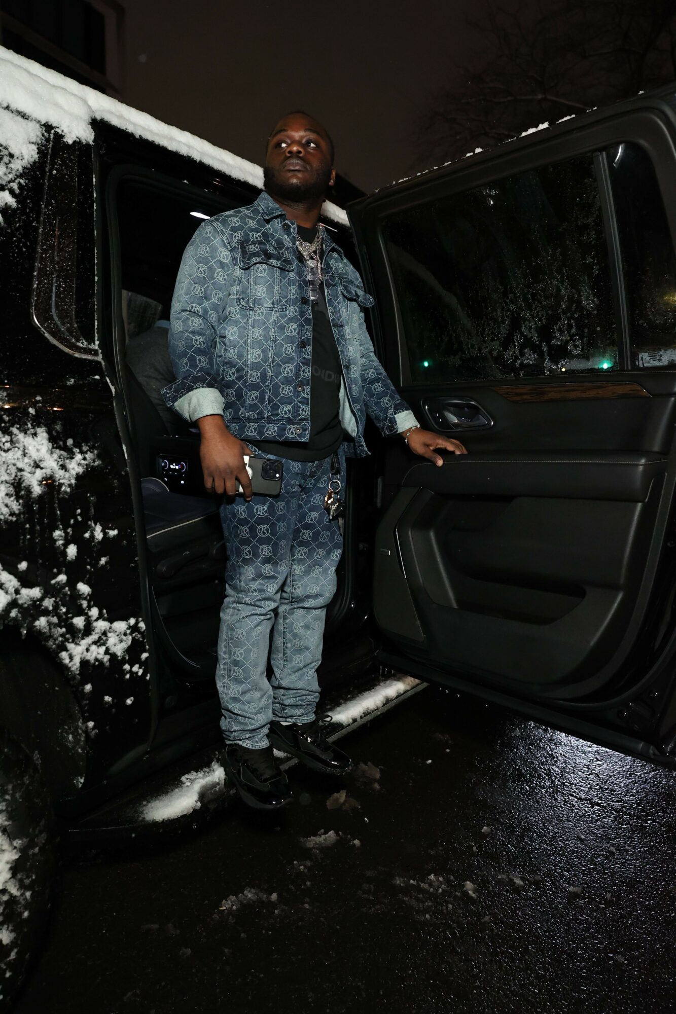 Man standing outside a vehicle at night, wearing a patterned shirt and jeans, with trees and a building in the background.