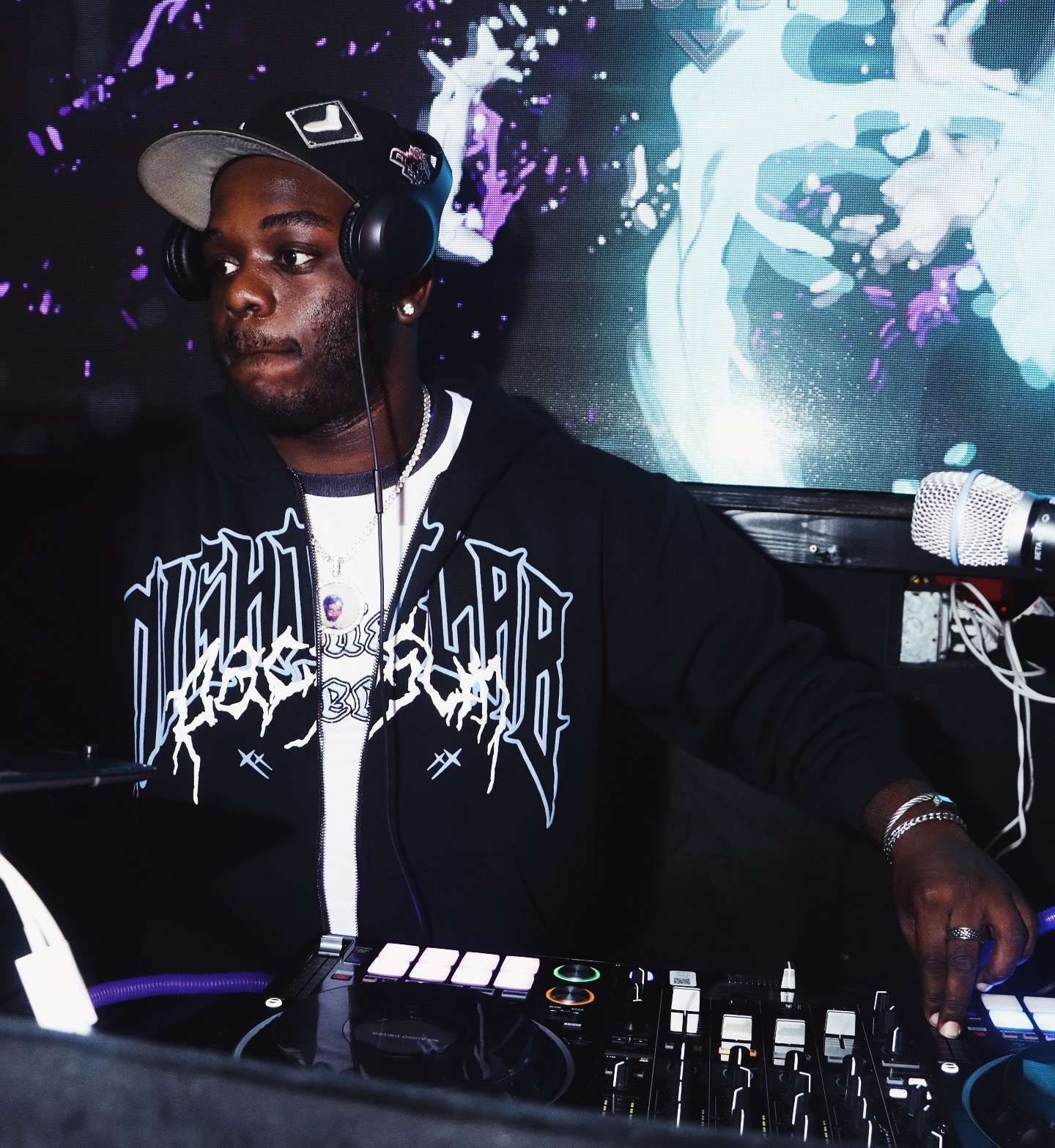 DJ wearing headphones and a cap, operating DJ equipment with a colorful digital background.