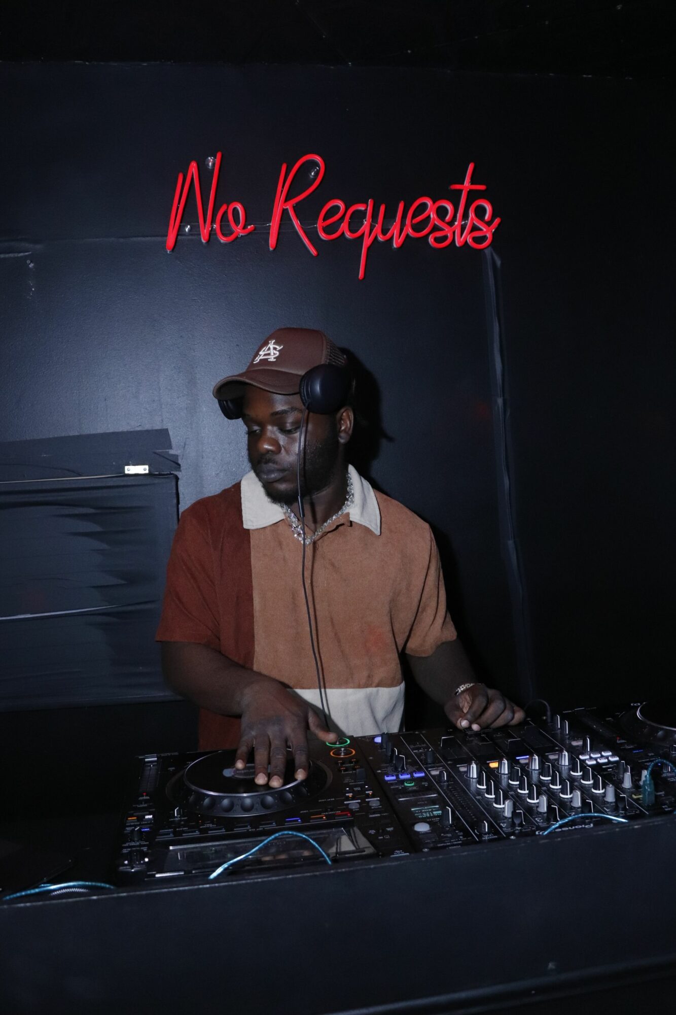 DJ wearing headphones and a brown jacket, standing behind a mixing console in a dark room with red text on the wall.