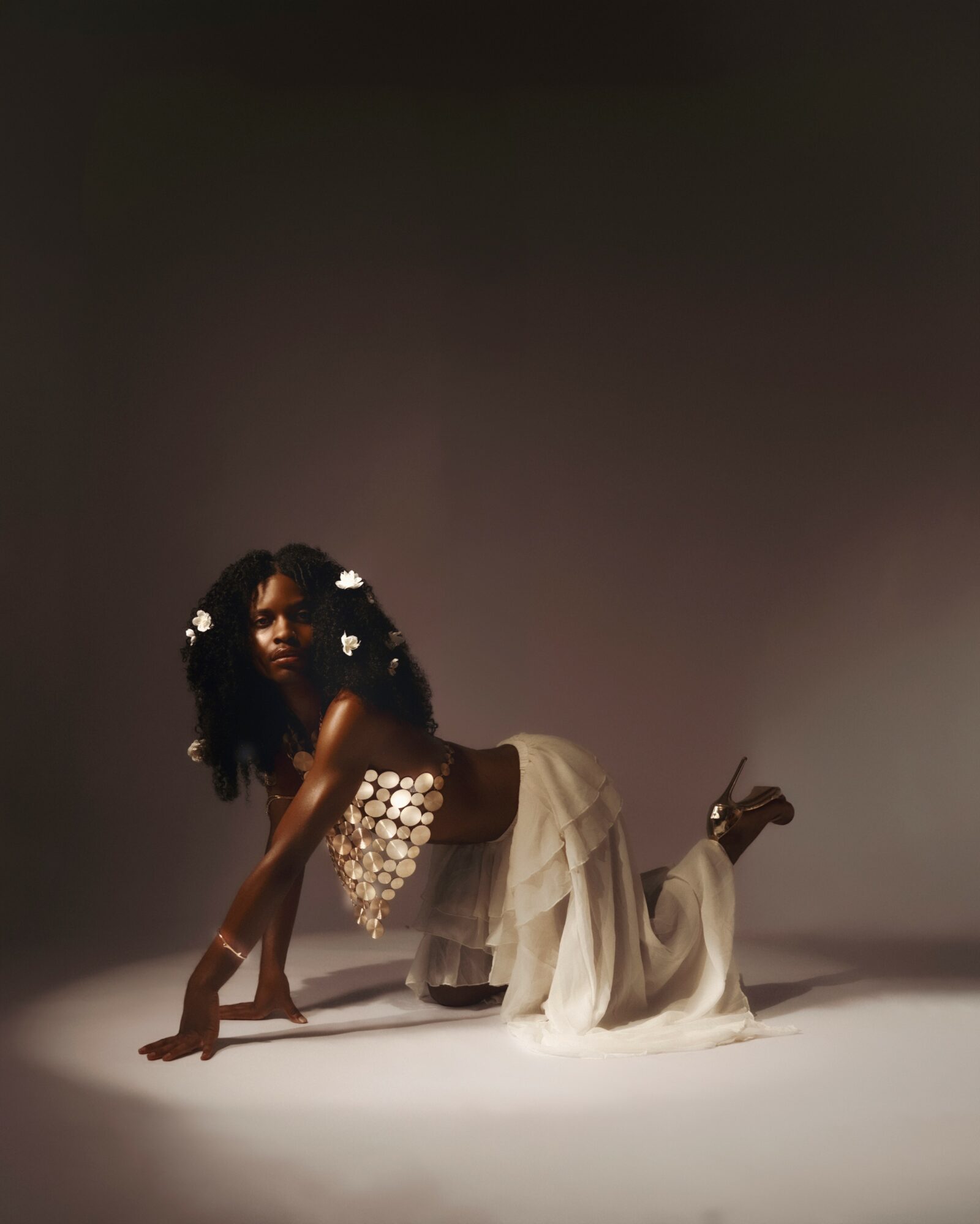 Person with curly hair and floral accessories kneels on the floor, wearing a white dress, holding a stick, in a studio.