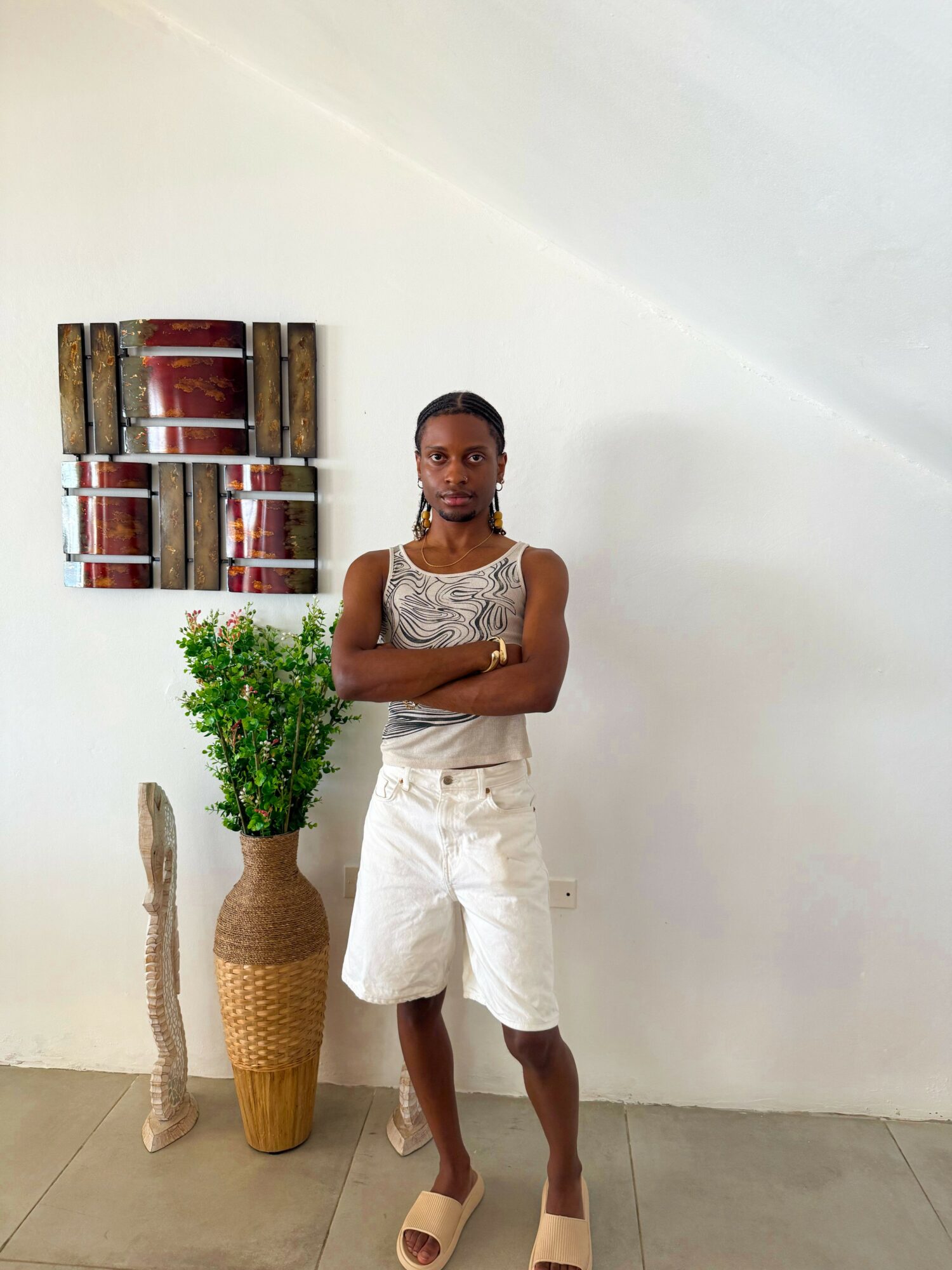 Woman standing with arms crossed in front of a white wall, next to a potted plant and decorative items.