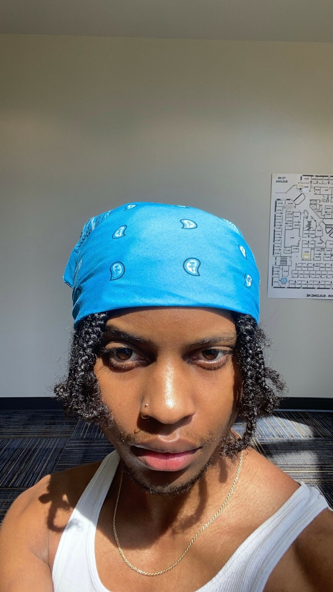 Person wearing a blue bandana with white patterns, looking directly at the camera, with a plain wall and chart in the background.