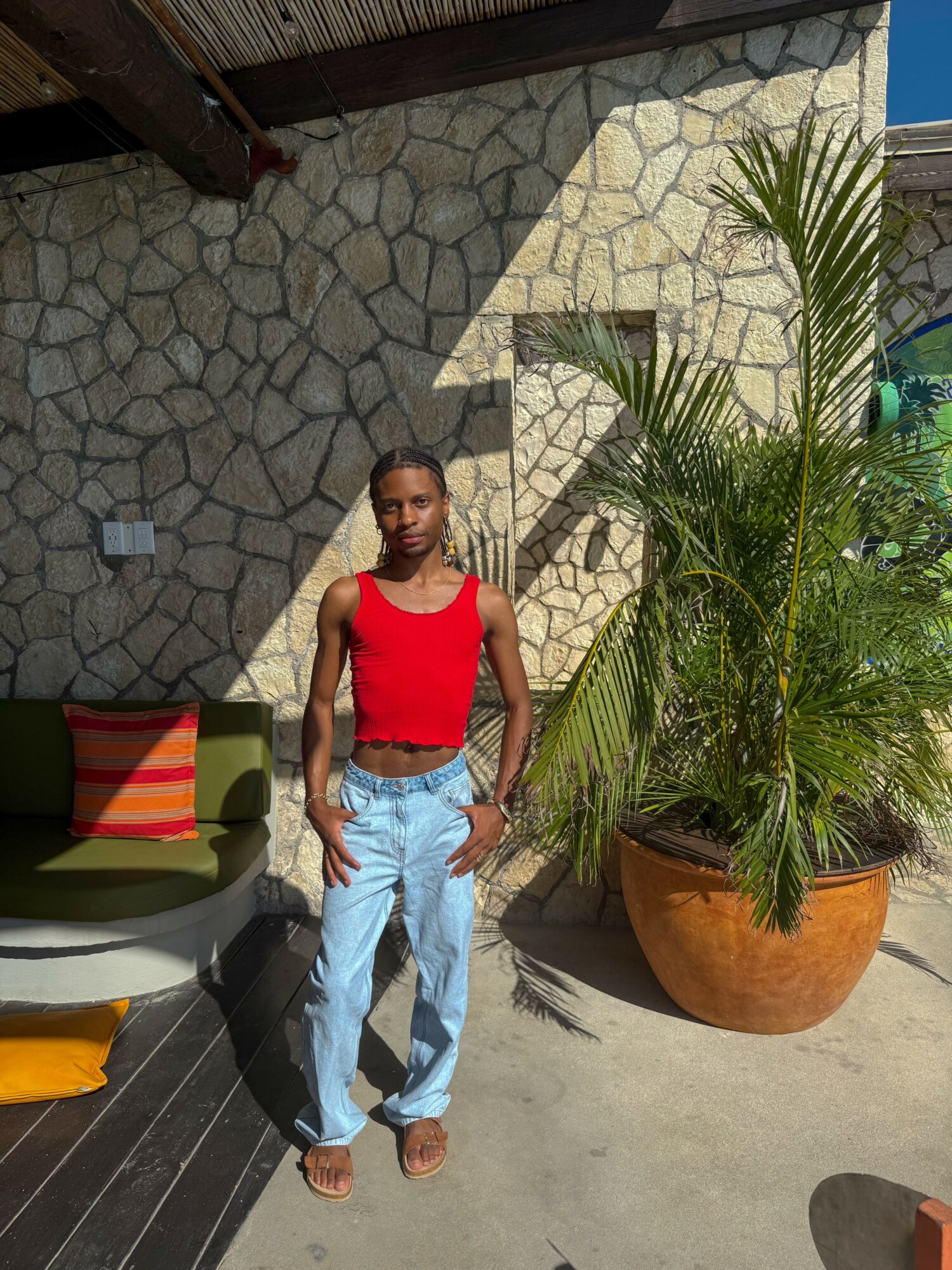 Young man standing outdoors next to a large potted plant, wearing a red tank top and light blue jeans.