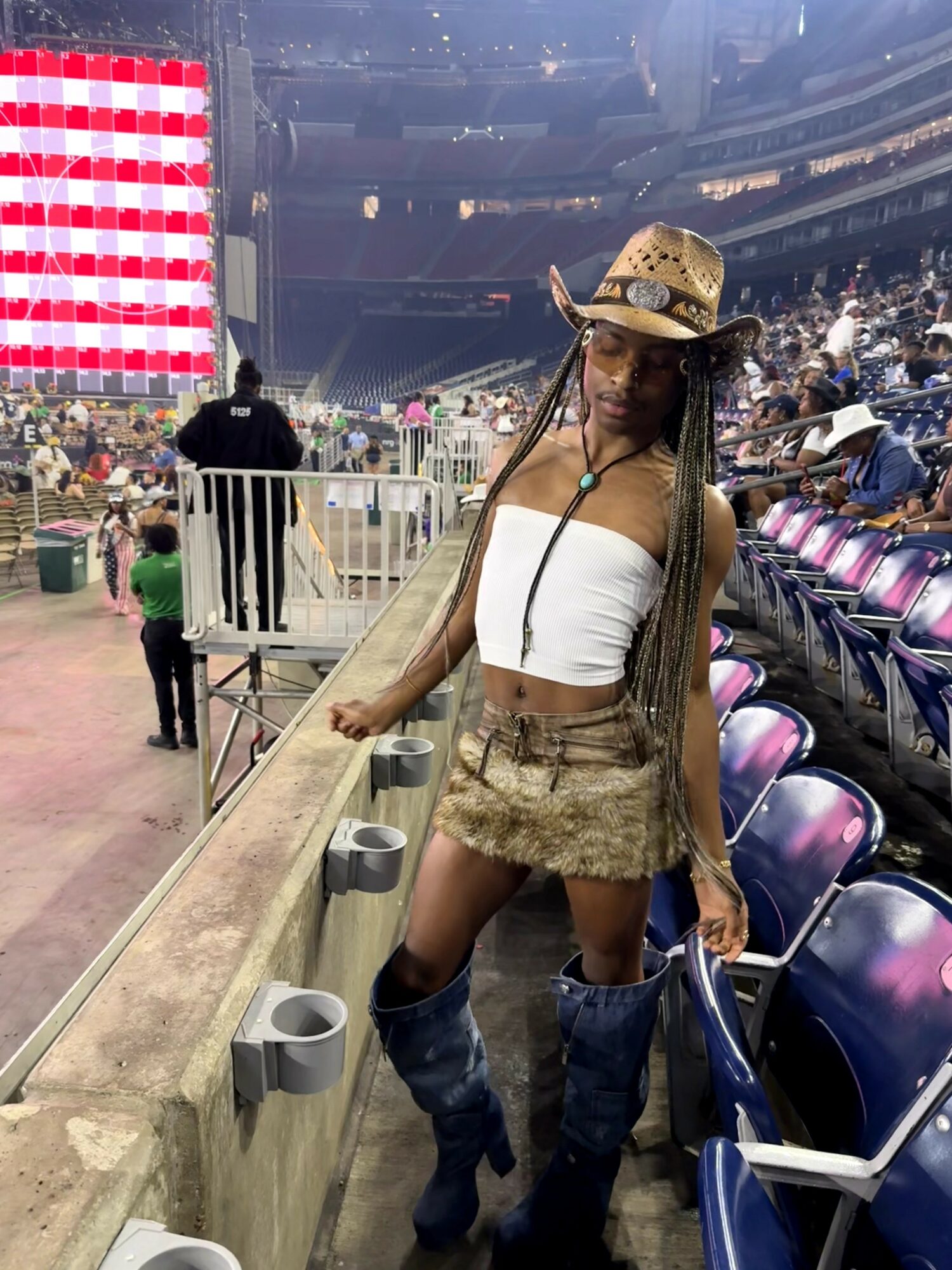 Young woman with long braids wearing a cowboy hat, white crop top, and shorts, standing at a stadium event.