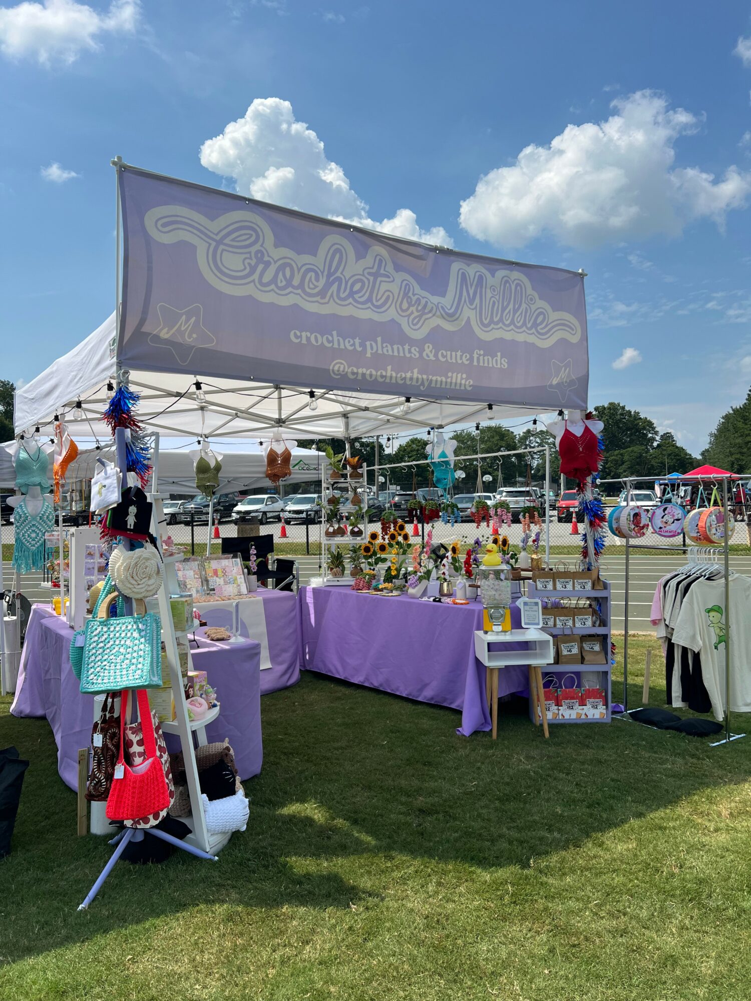 Outdoor craft booth with tables displaying handmade items, under a large sign, on a grassy area with blue sky and clouds.