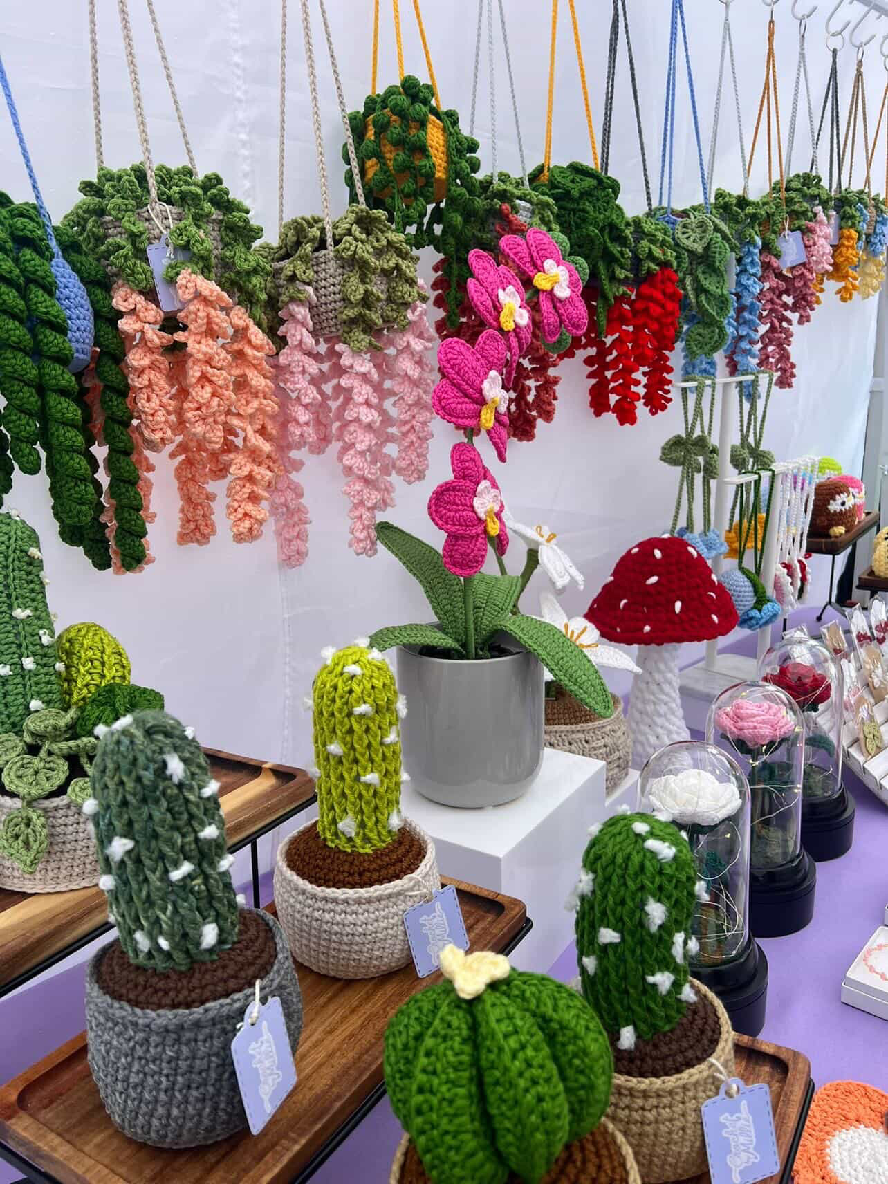 Colorful hanging and potted yarn or fabric cacti and flowers display on a table, with a white background.