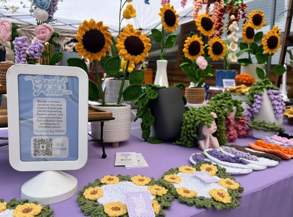 Display of sunflower and floral arrangements on a purple tablecloth with informational sign and decorative items.