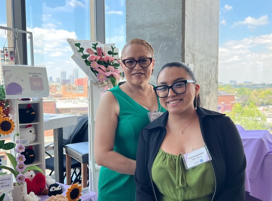 Two women smiling indoors with a city view outside window, one wearing glasses and green dress, the other in black jacket and green top.