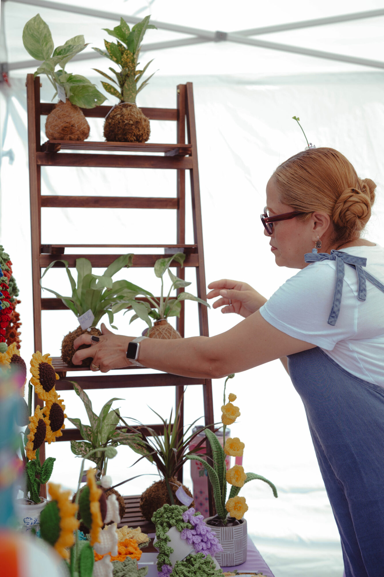 Woman tending to potted plants on a wooden ladder display, with flowers and greenery around her, indoors or at a market.