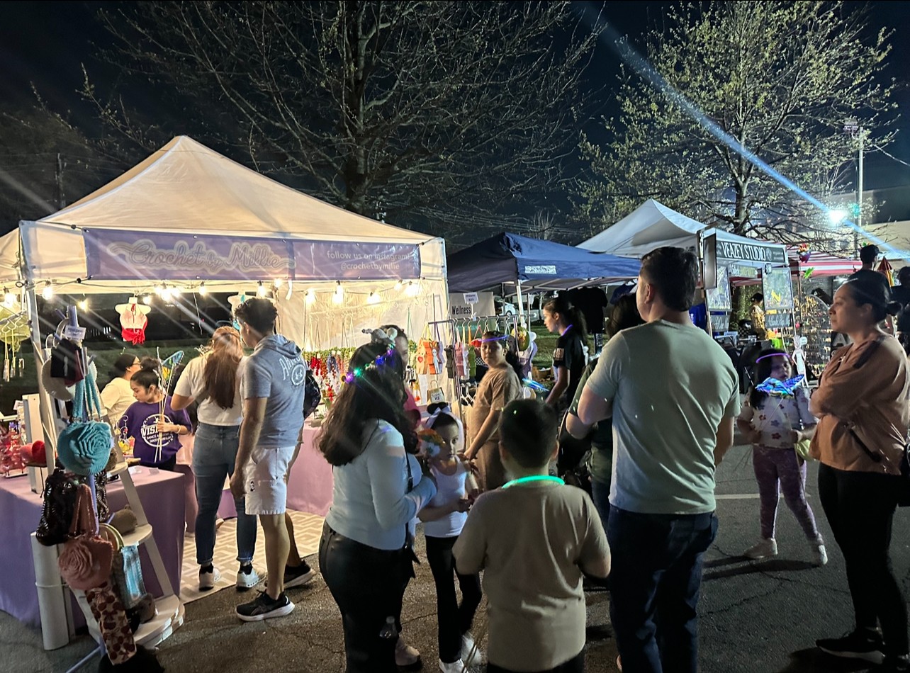 Night scene with tents, people, and trees, illuminated by lights at an outdoor event.