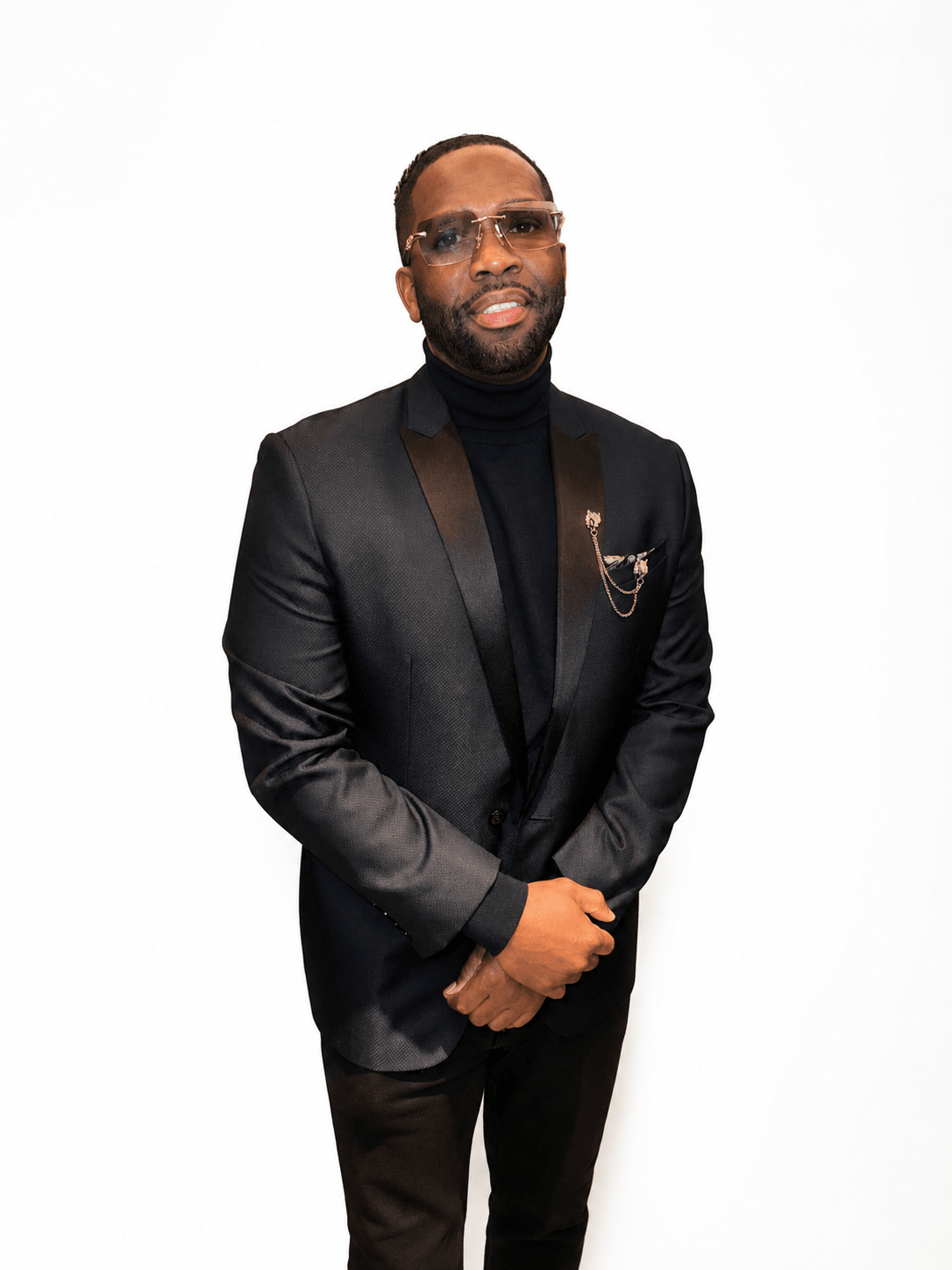 Man in black suit with glasses and a chain, standing against a white background.