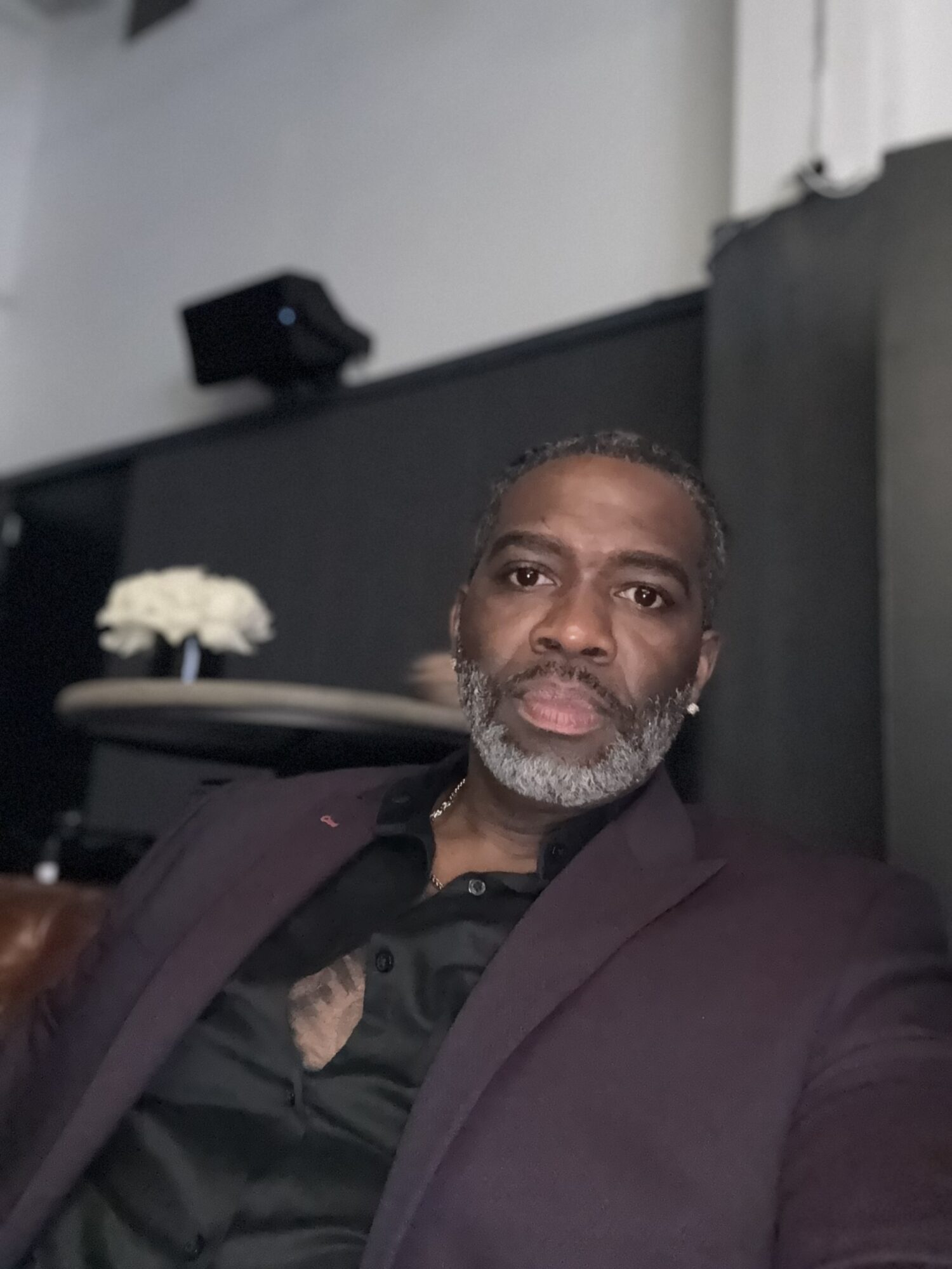 Man with gray beard and short hair wearing a dark blazer and black shirt, sitting indoors.