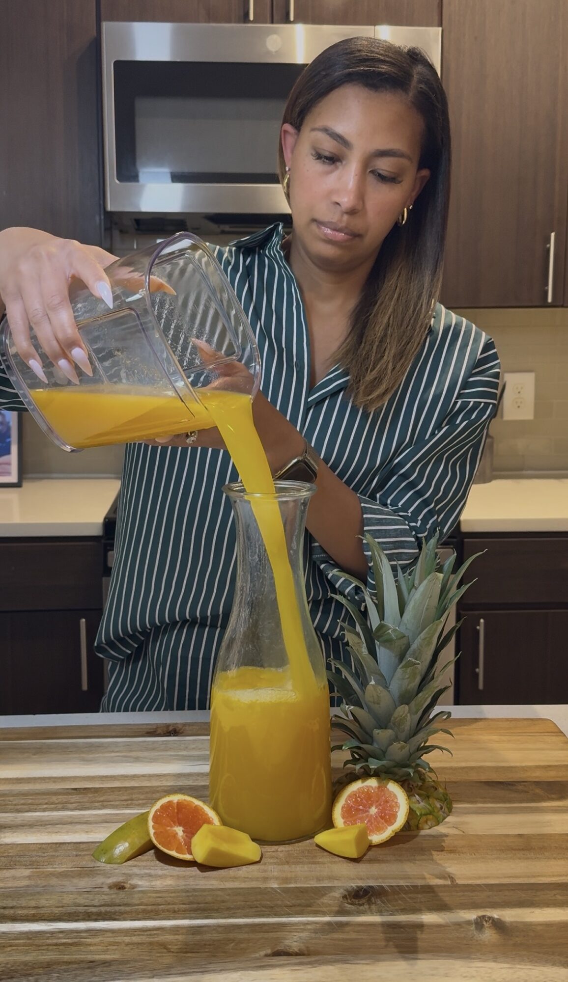 Woman pouring yellow juice into a glass with pineapple and citrus fruits on a kitchen counter.