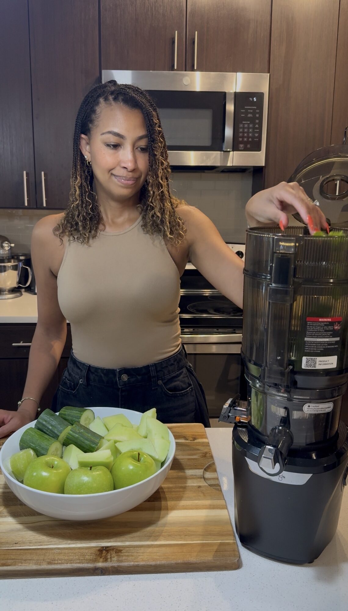 Woman in a kitchen with a bowl of green apples and a juice machine, smiling and preparing to make juice.