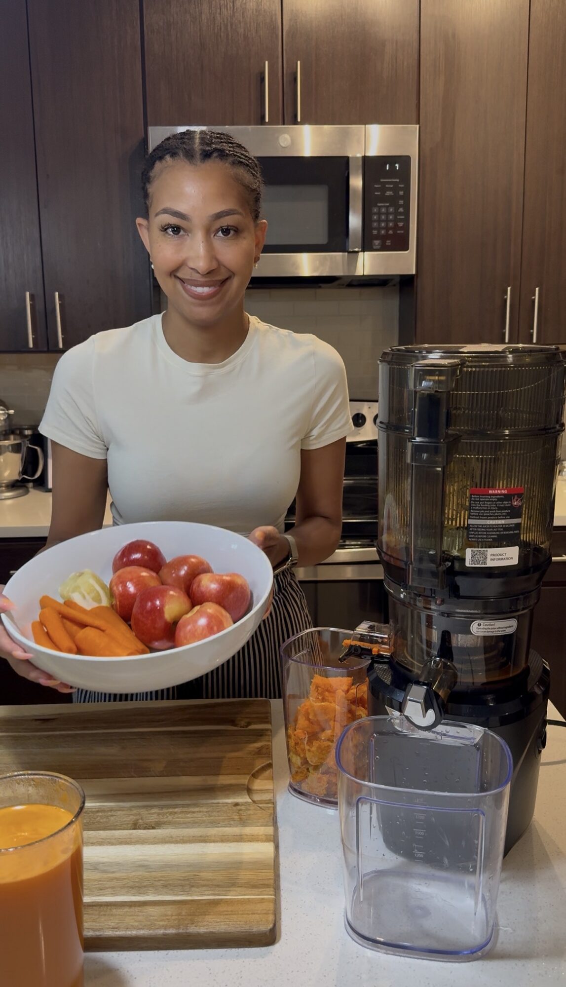 Smiling woman standing in kitchen with bowl of apples and carrots, blender, and glass of orange juice in foreground.