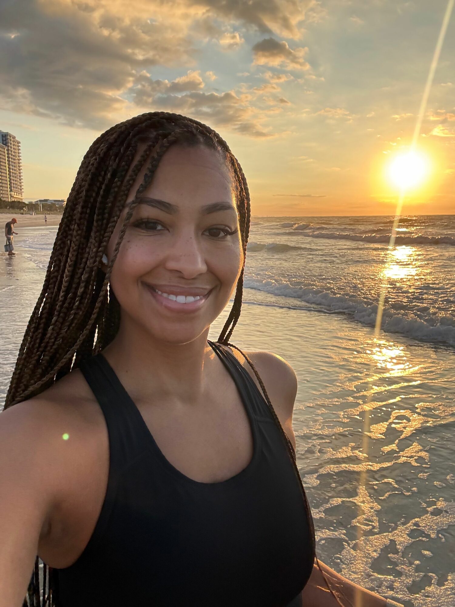 Smiling woman with braided hair takes a selfie at sunset on the beach, ocean waves in background.