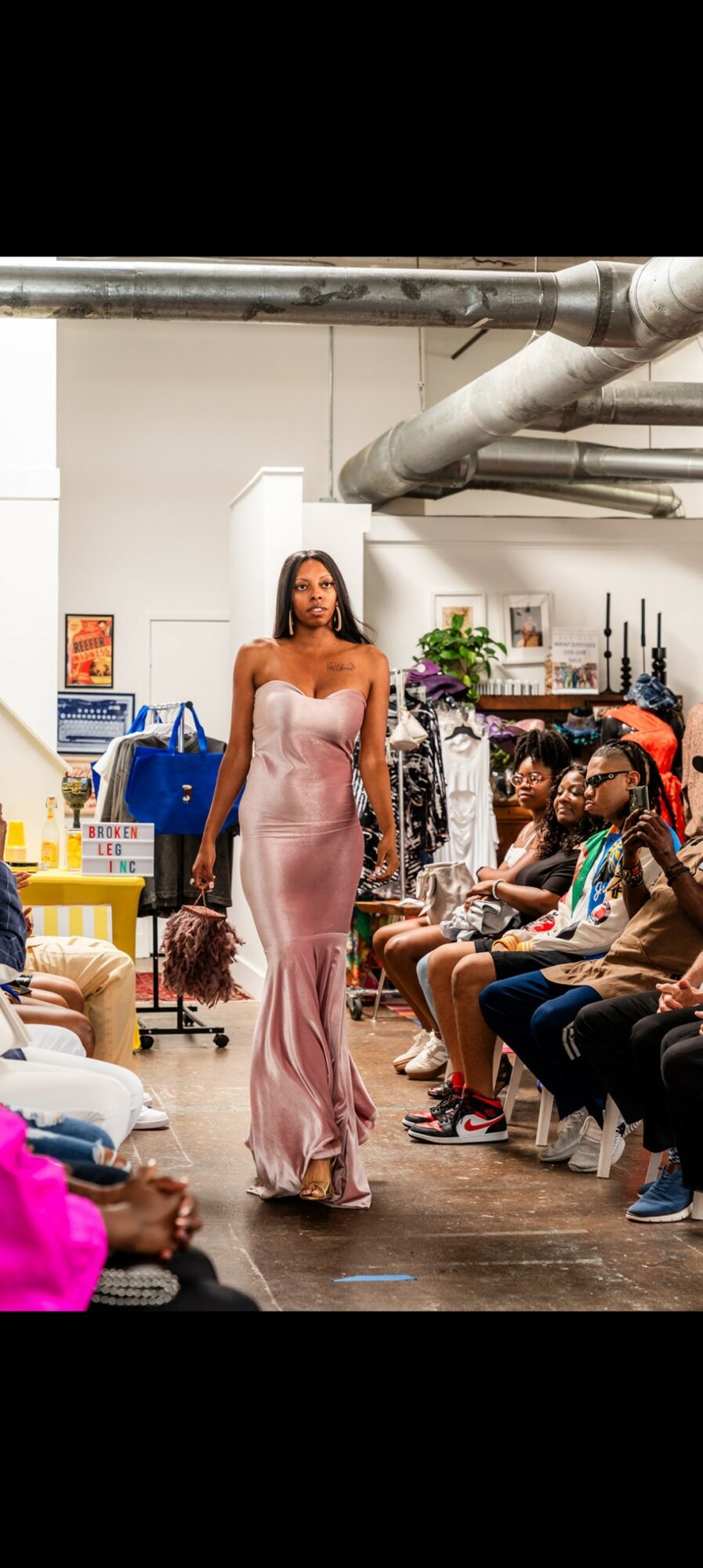 Model walking down runway in pink dress, audience seated on both sides, industrial background with pipes and shelves.